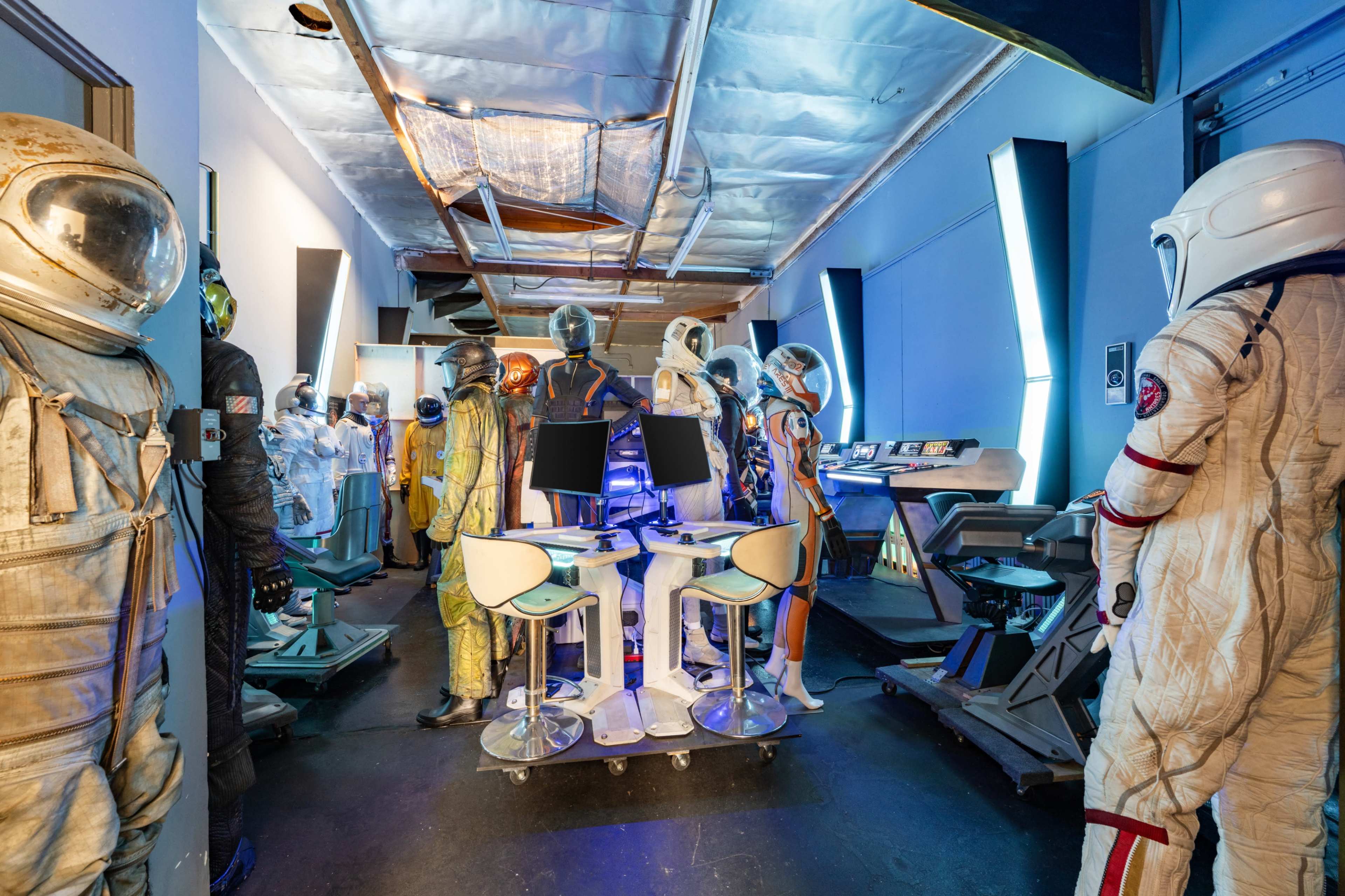 The image shows a room filled with various astronaut suits and futuristic equipment, illuminated by blue lighting.