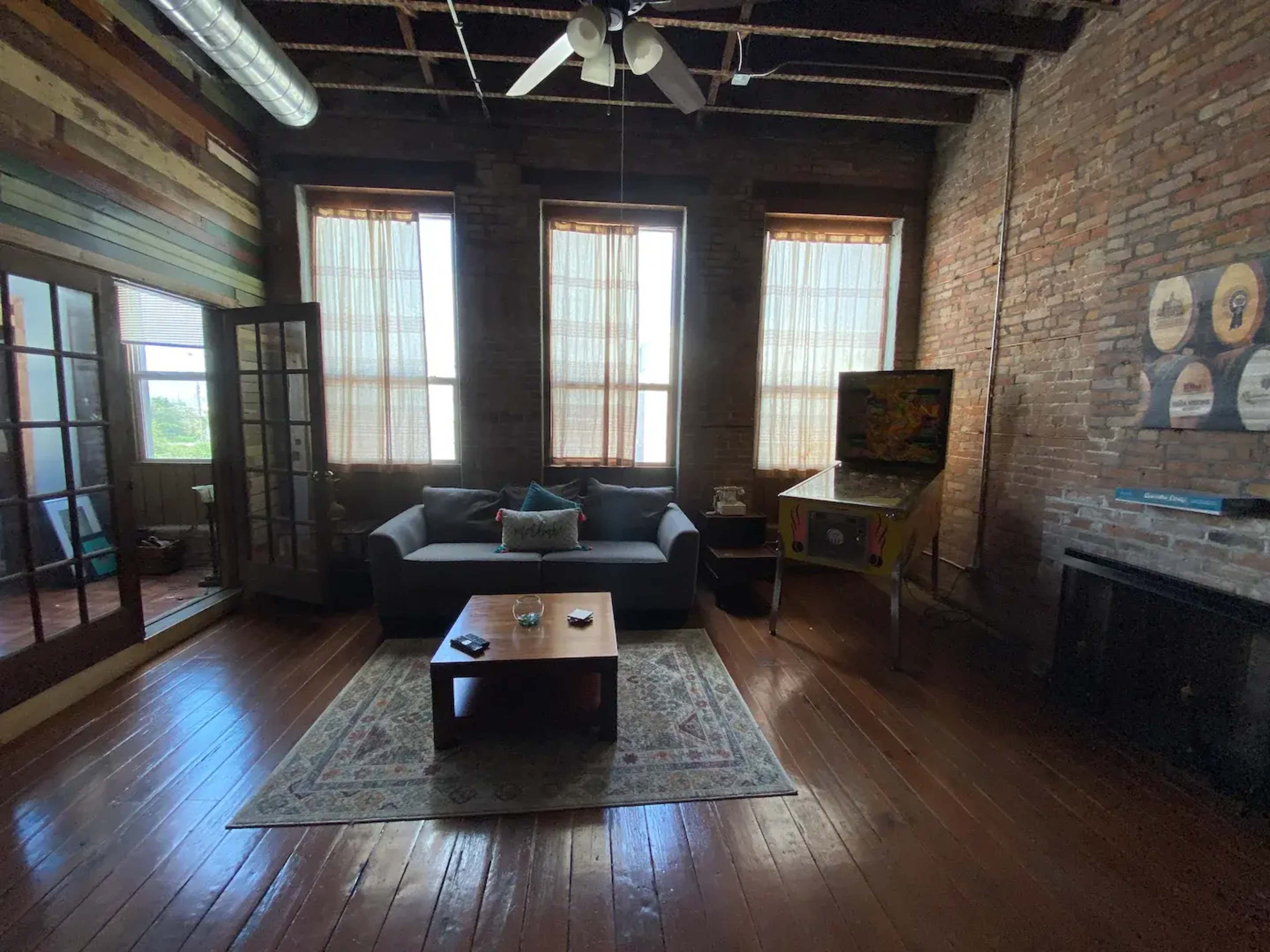 The image shows a living room with exposed brick walls, large windows adorned with sheer curtains, a dark sofa, a coffee table on a rug, and a vintage pinball machine in the corner.