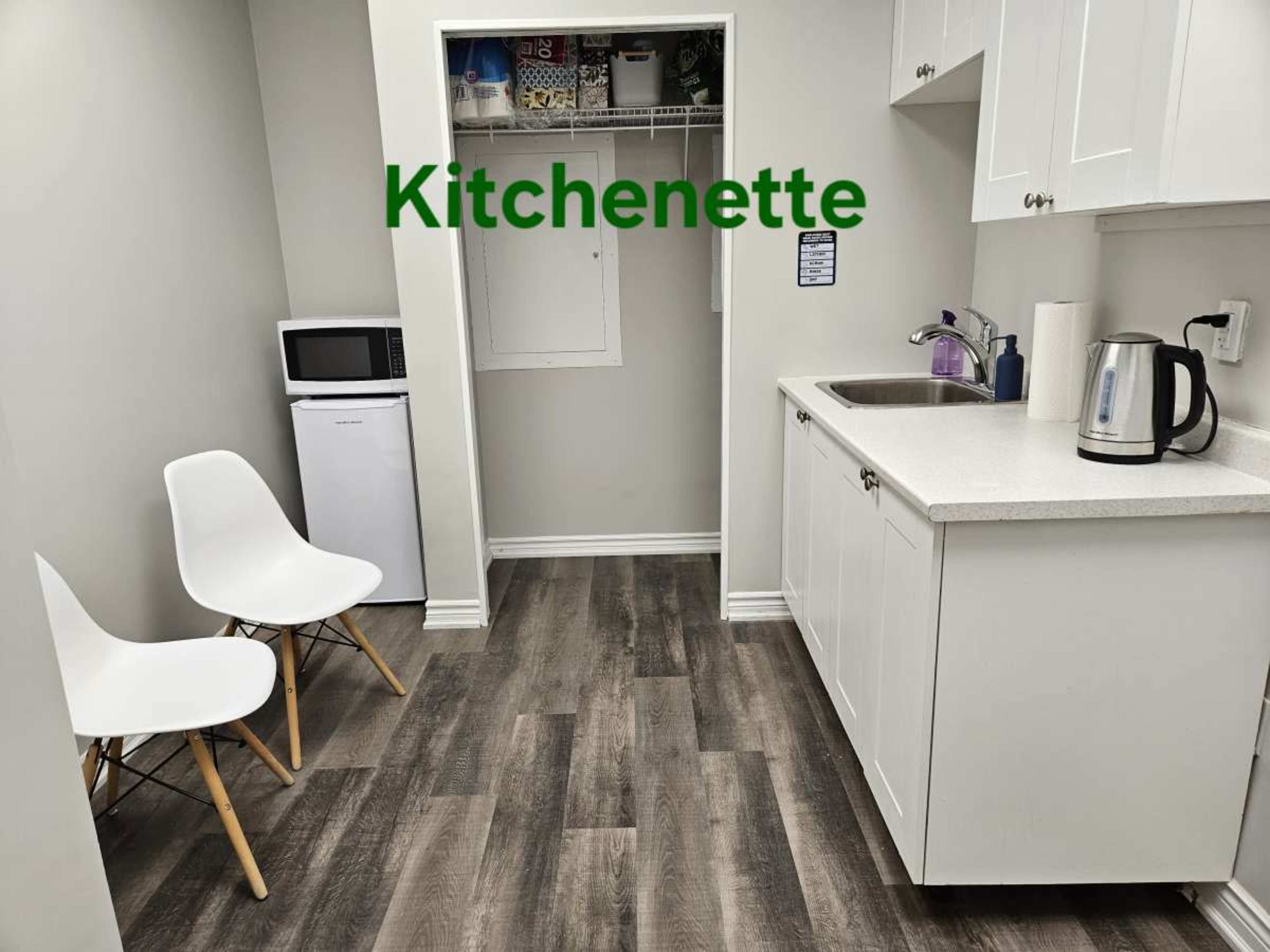 A small kitchenette features a microwave, a mini-fridge, a sink, a kettle, and two white chairs against a light gray wall.