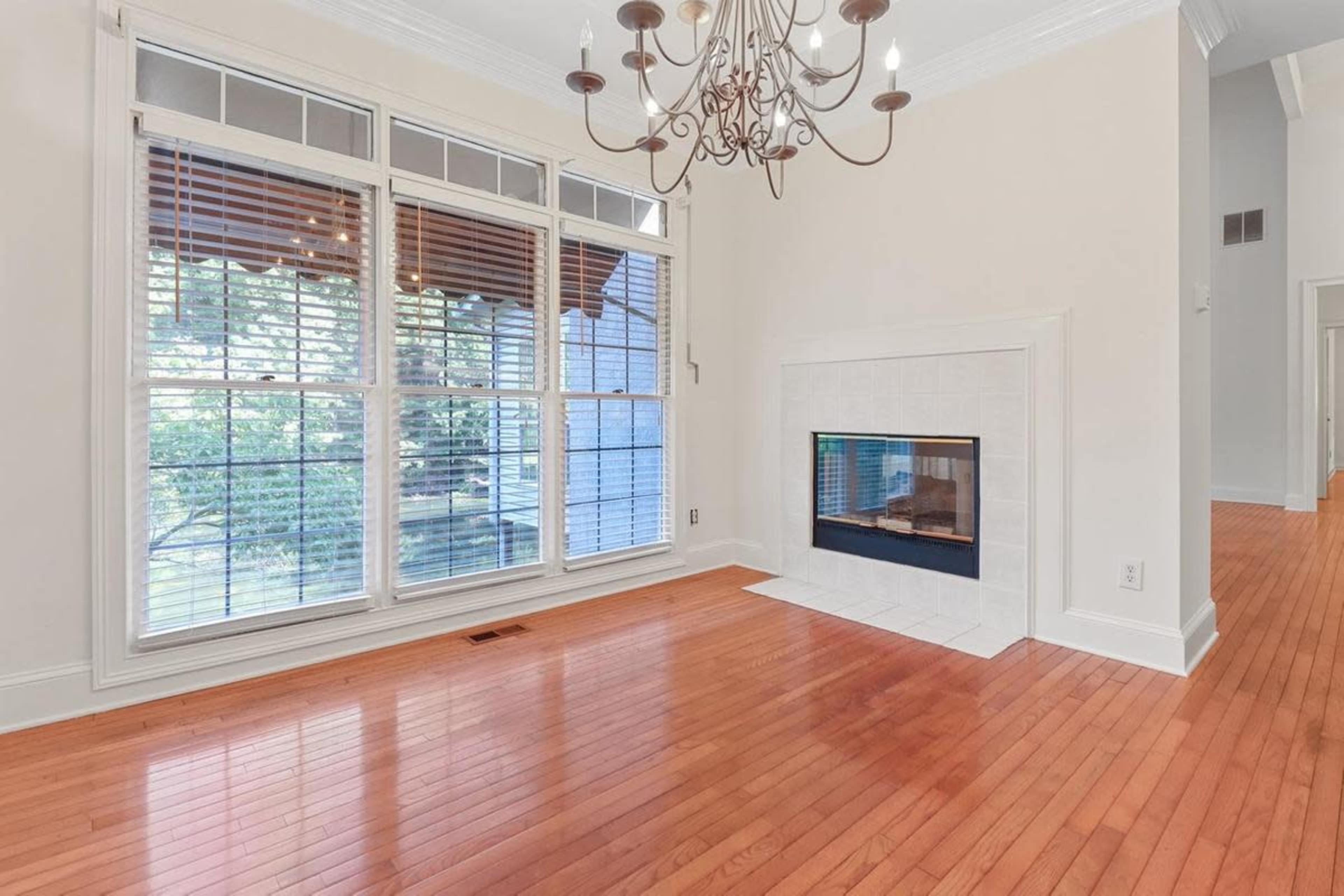 The image shows a spacious room with hardwood floors, large windows with blinds, a fireplace integrated into the wall, and a chandelier hanging from the ceiling.