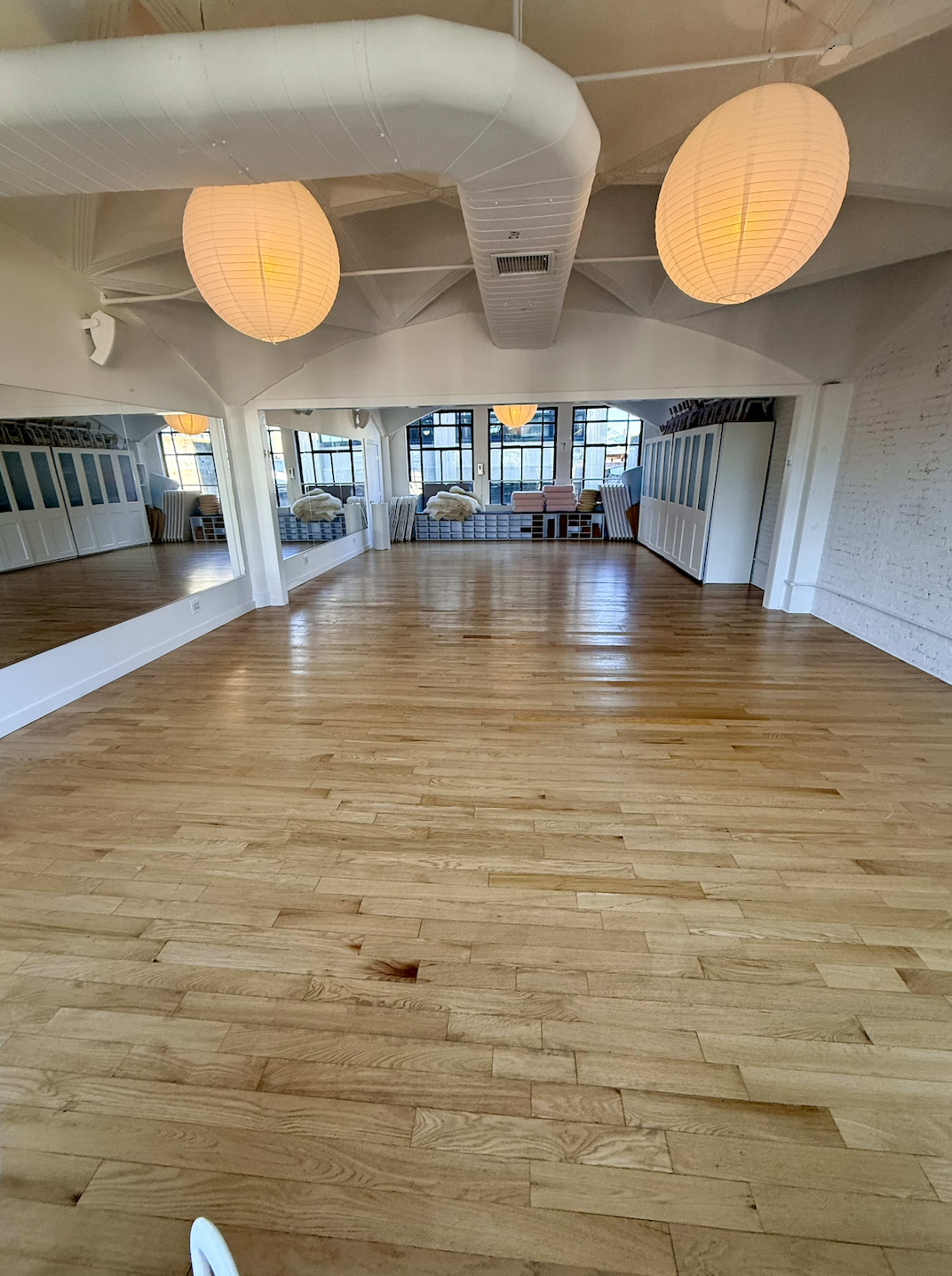 The image shows a spacious, well-lit studio with wooden flooring, large windows, and two hanging light fixtures.