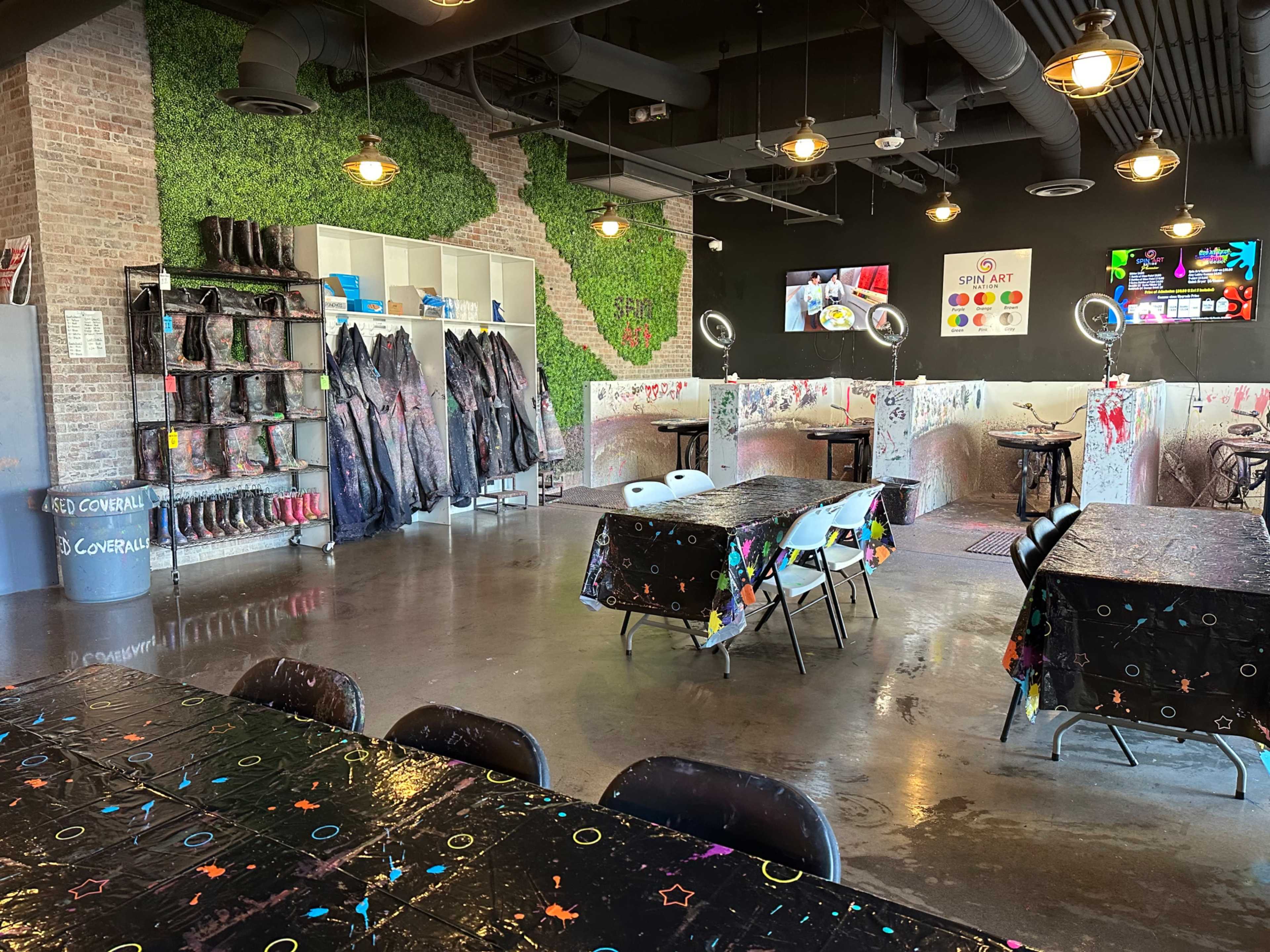 An art studio with tables covered in colorful tablecloths, black chairs, and a wall decorated with green foliage, alongside shelves of paint supplies and protective gear.