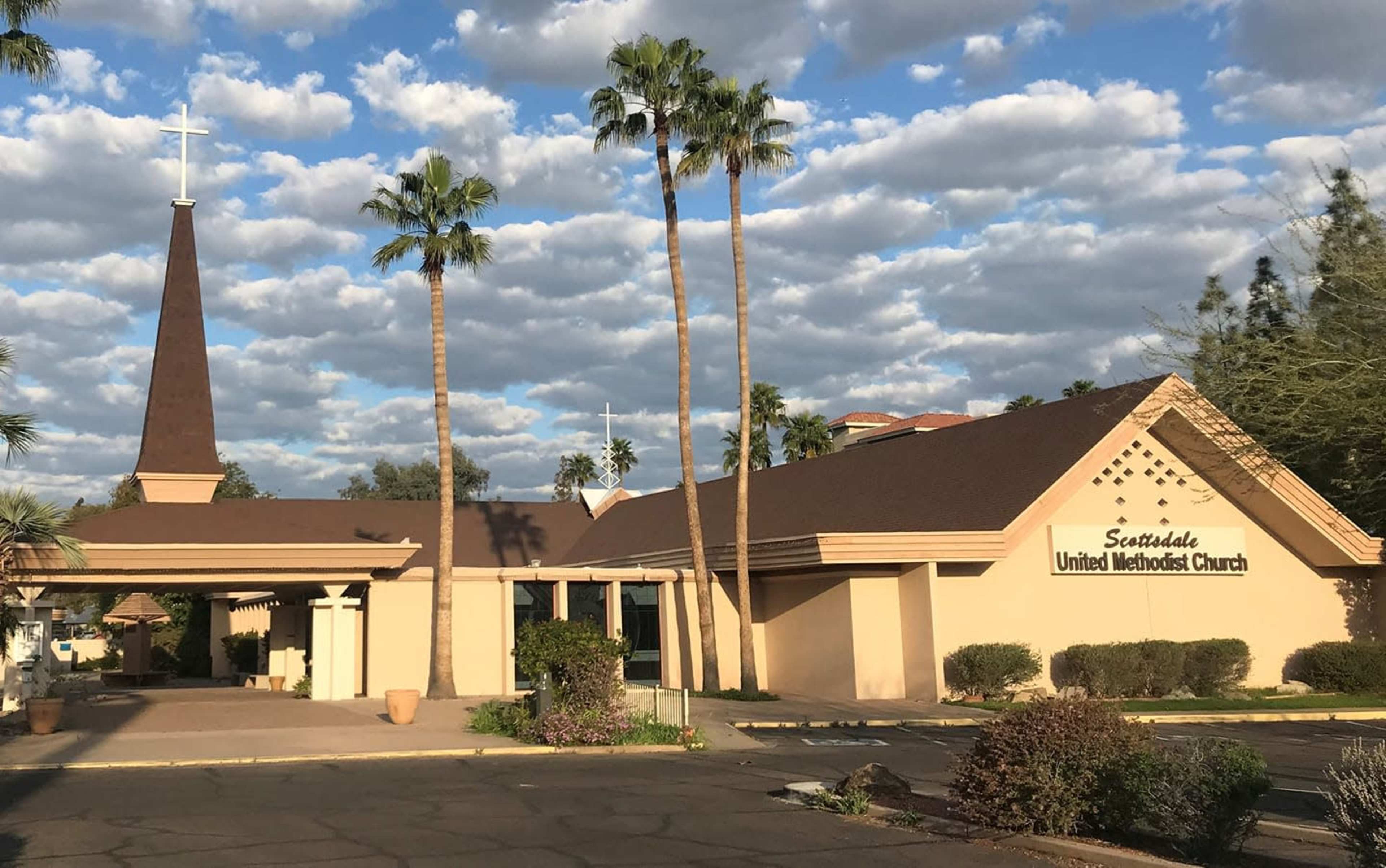 The image shows Scottsdale United Methodist Church, featuring a tall spire and palm trees under a partly cloudy sky.
