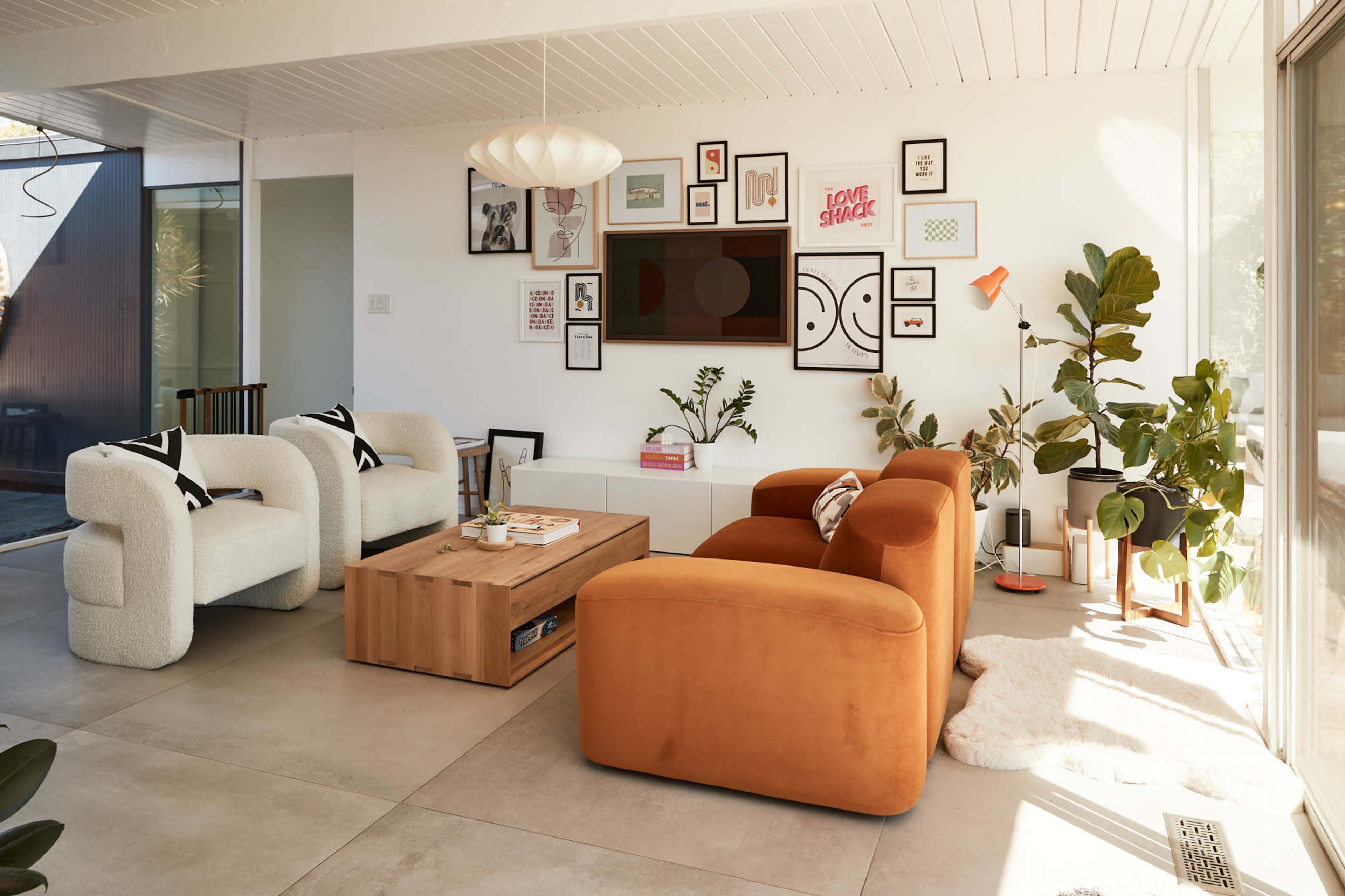 A modern living room features a mix of white and orange furniture, a wooden coffee table, framed artwork on the walls, and several indoor plants.