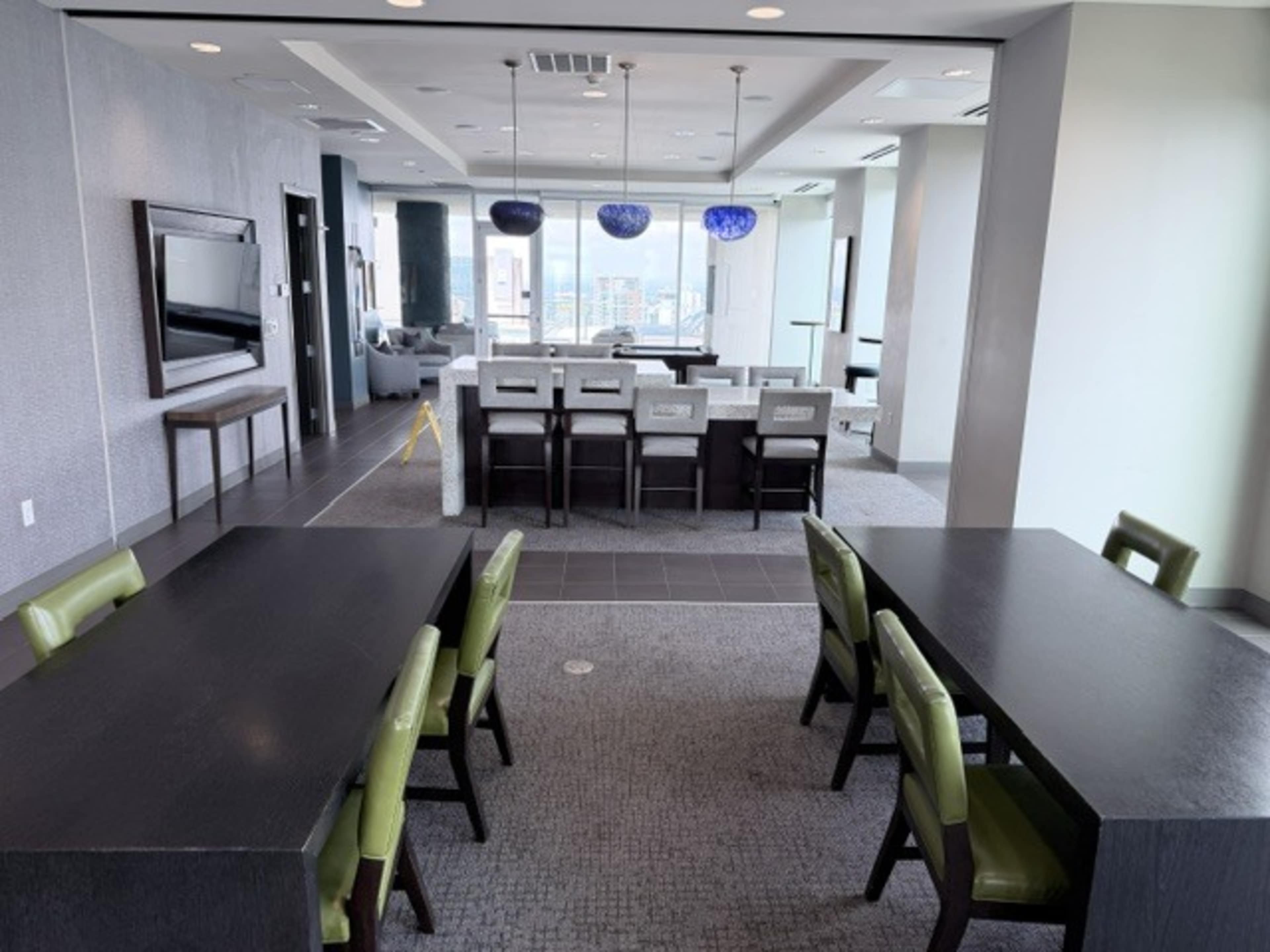 The image shows a modern, spacious room with two dark tables and green chairs, leading to a dining area with gray upholstered seating and decorative pendant lights.