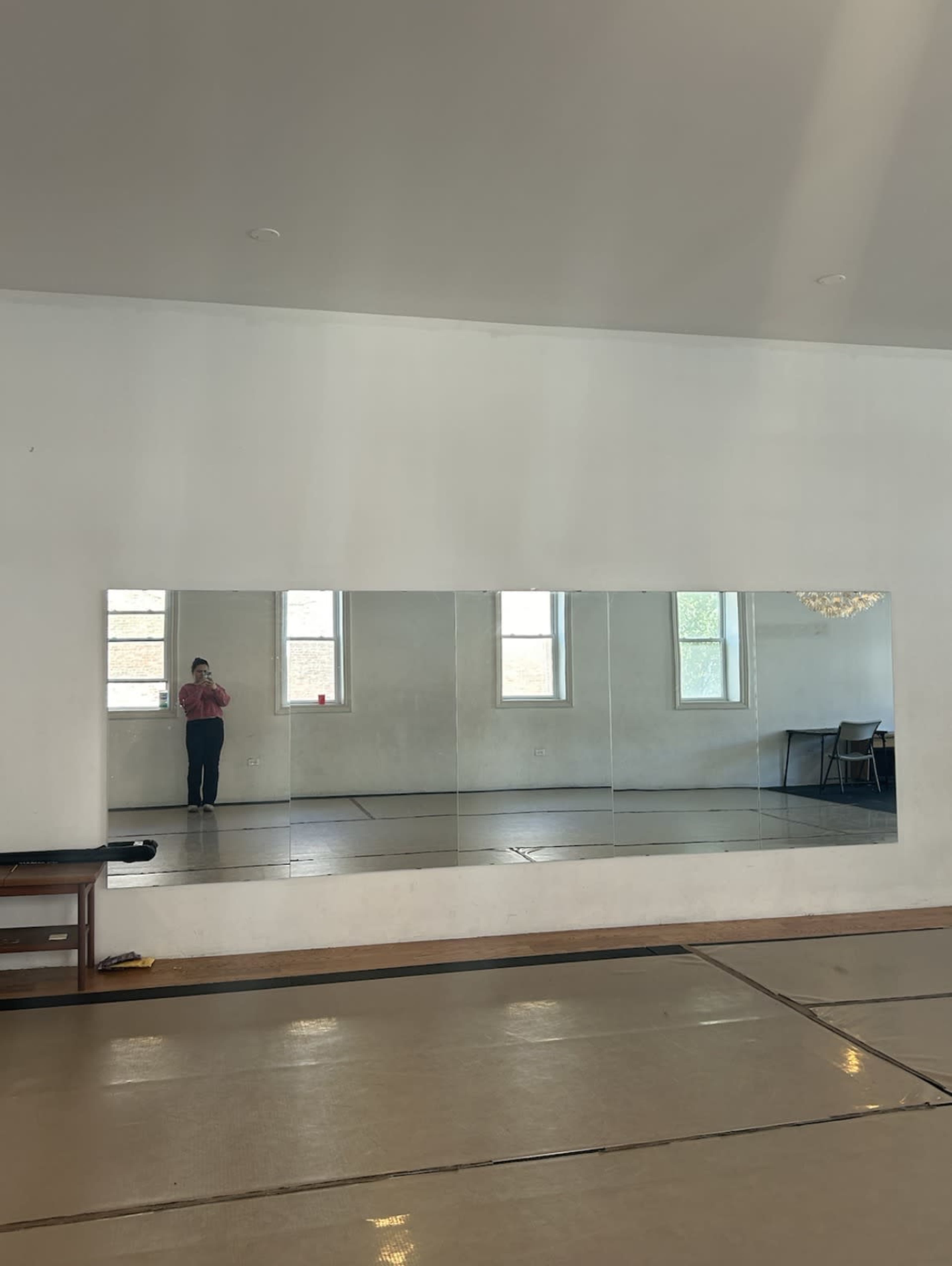 The image shows a spacious room with wooden flooring and large mirrors on one wall, reflecting windows and a person standing in front of them.