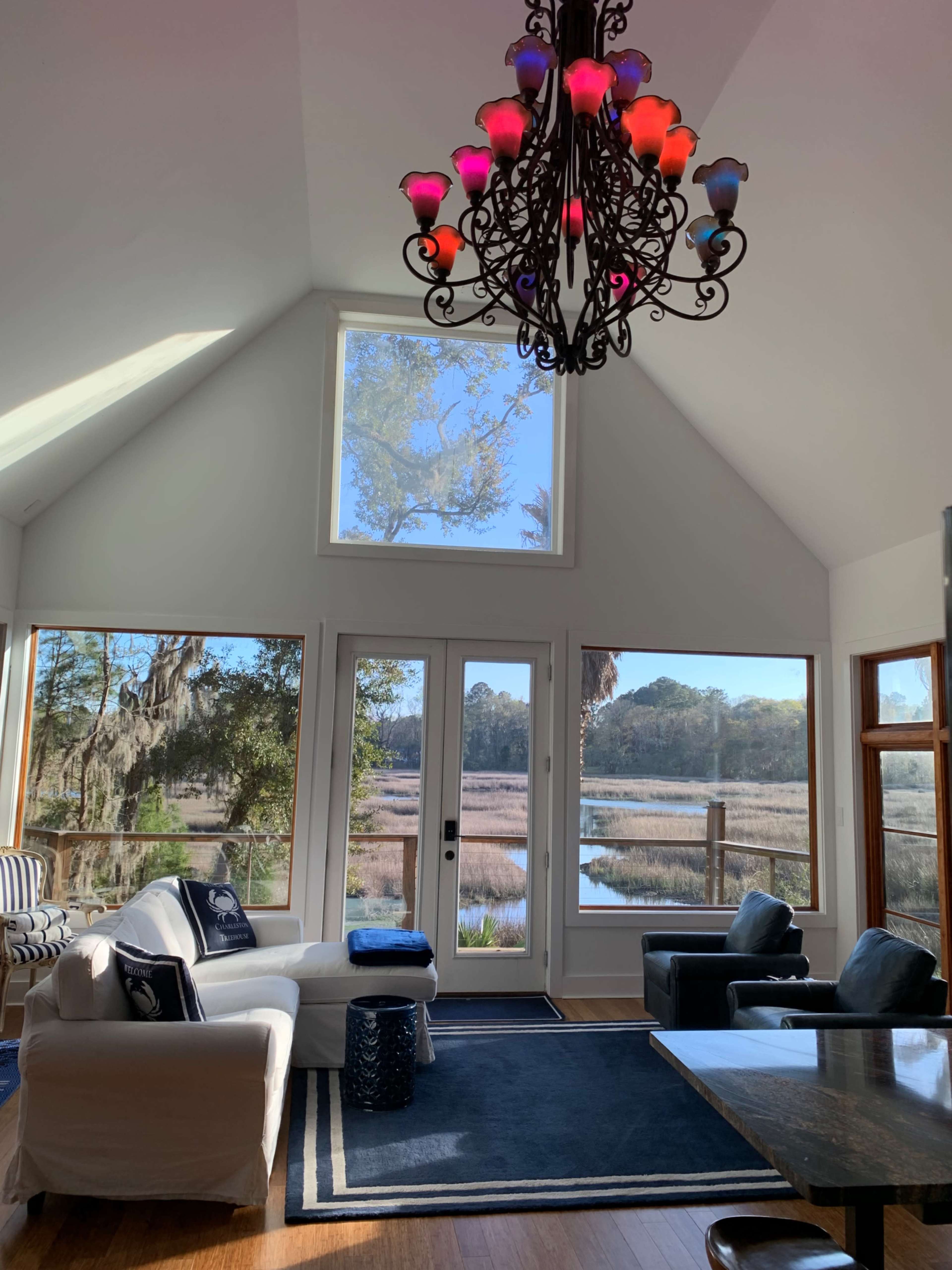 A bright, airy living room features large windows overlooking a marsh area, with a decorative chandelier hanging from a vaulted ceiling.