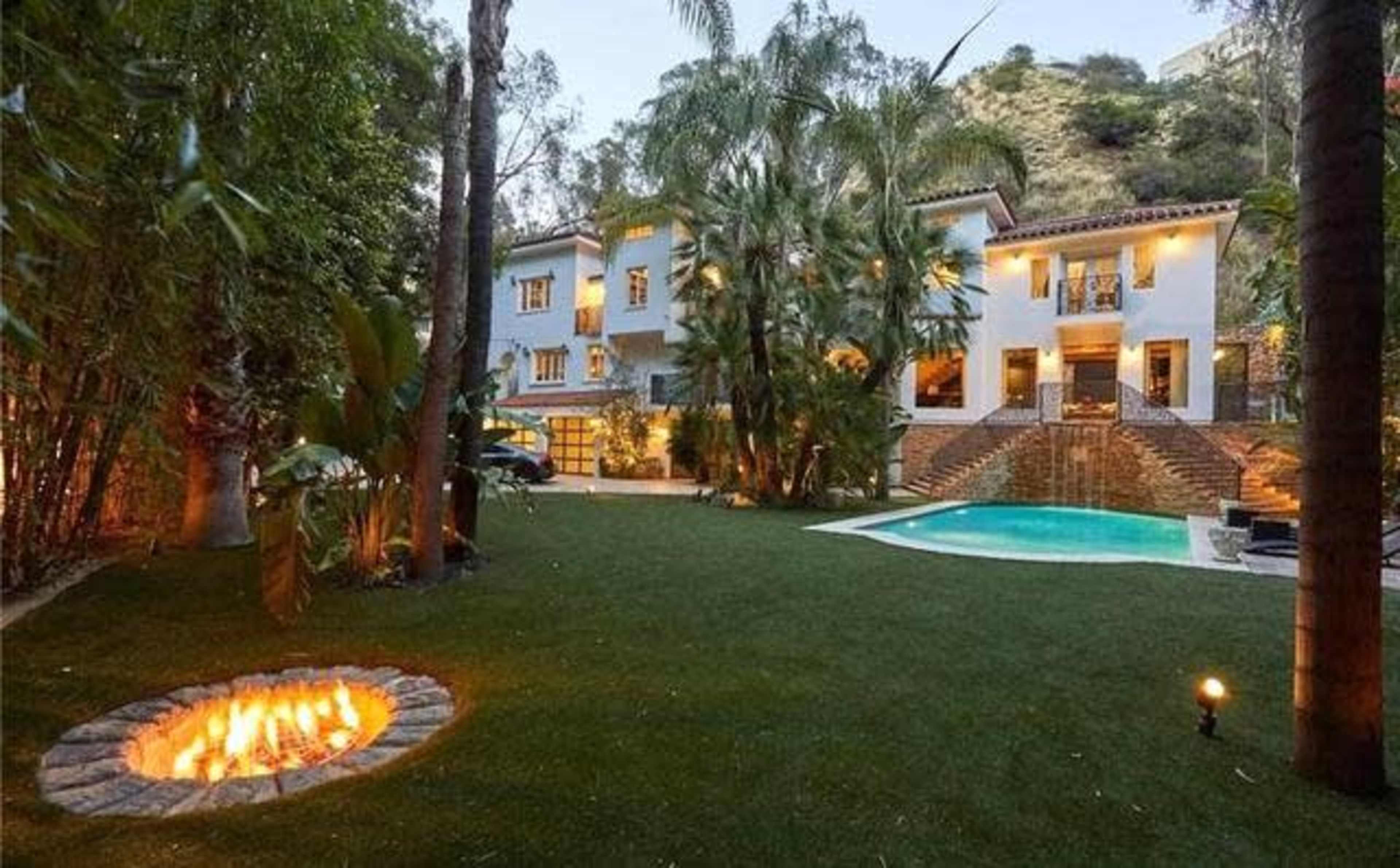 A two-story house surrounded by palm trees and a manicured lawn, featuring a pool and a fire pit in the backyard.