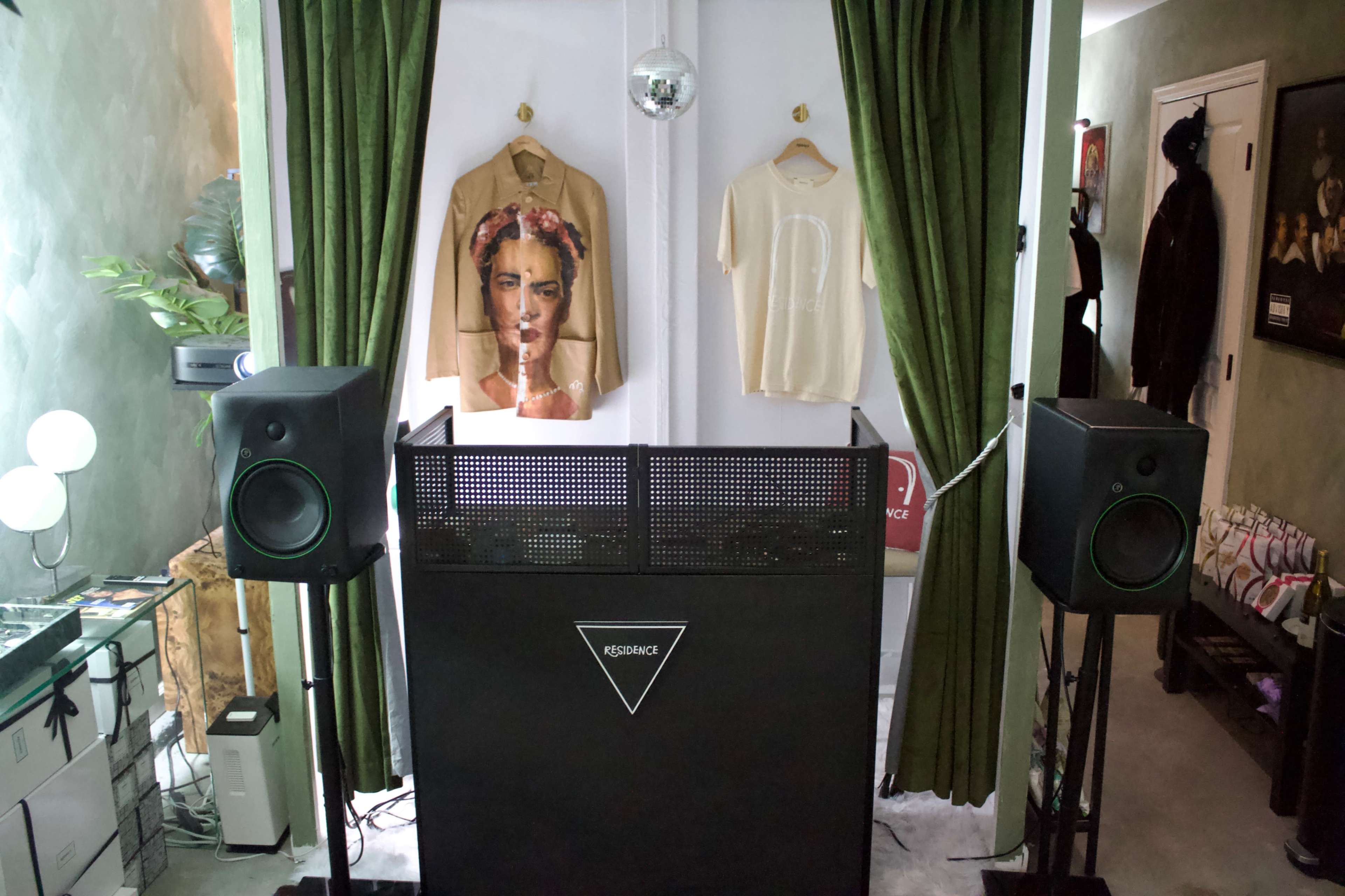 The image features a minimalist DJ setup with a black booth, two speaker systems, a disco ball hanging above, and a framed artwork of a woman on the wall.