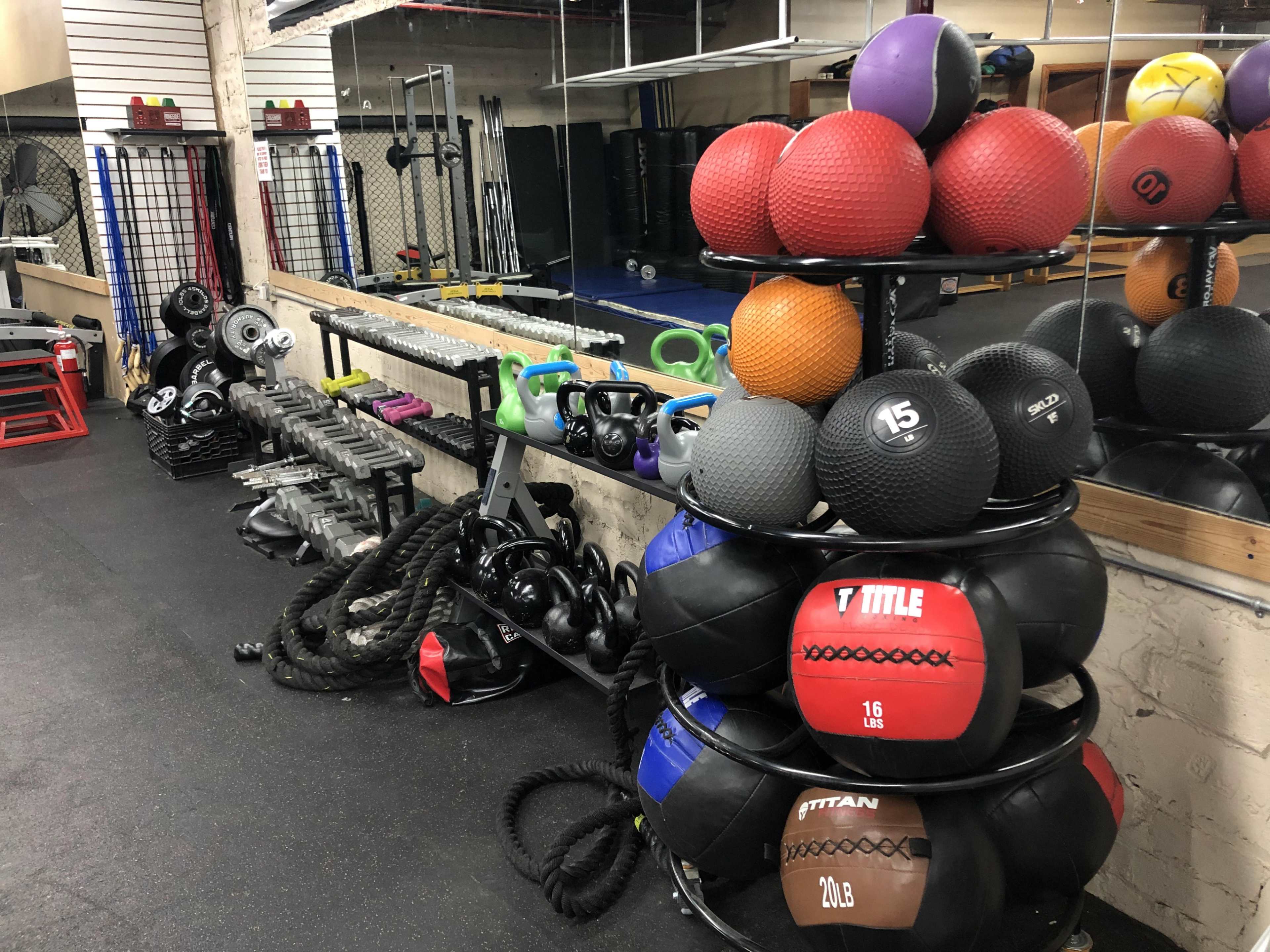 The image depicts a well-equipped gym space featuring stacks of medicine balls, various weights, dumbbells, and exercise equipment against a mirrored wall.