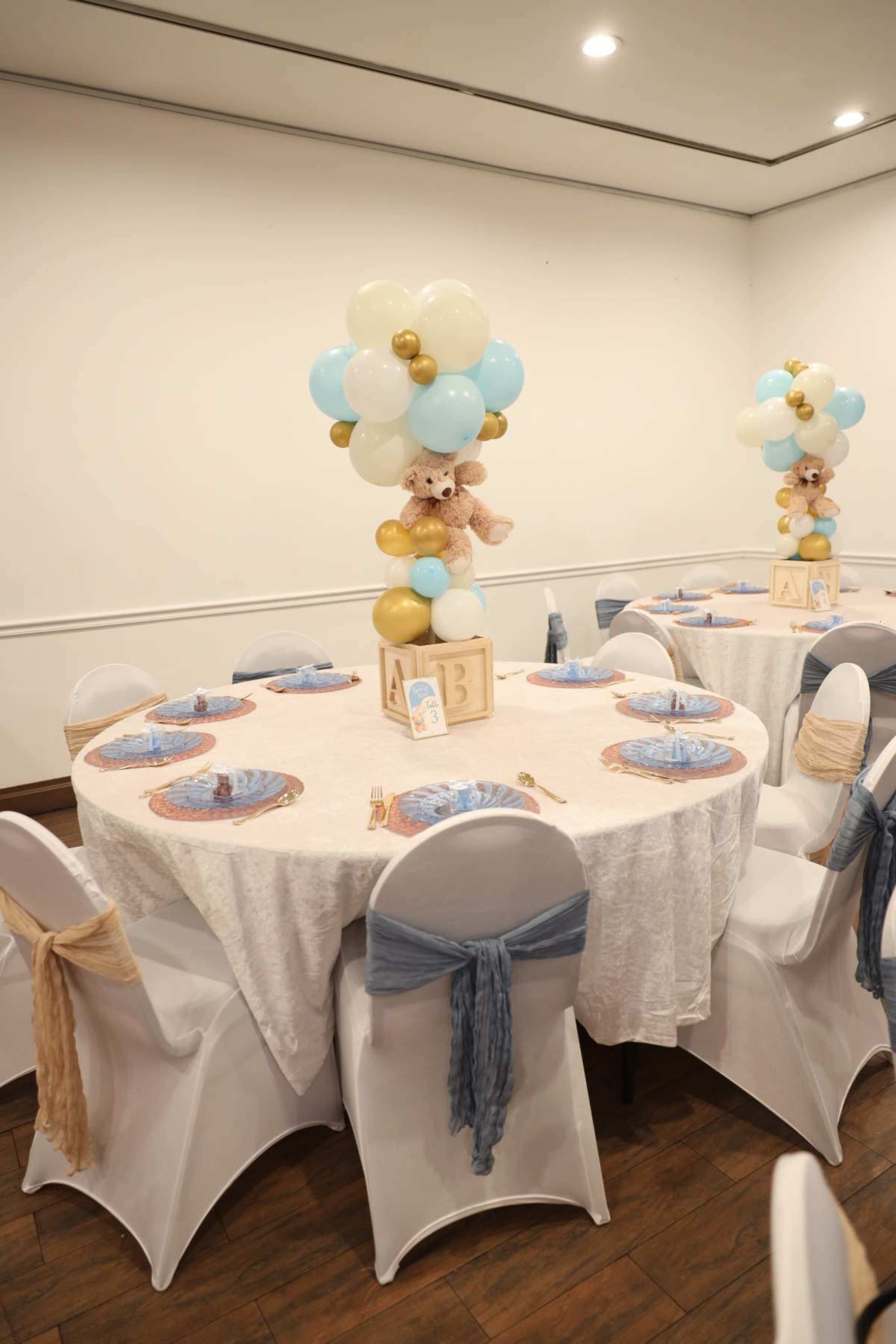 Two round tables are decorated with white tablecloths and playful centerpieces of balloons and teddy bears, arranged for a celebration.