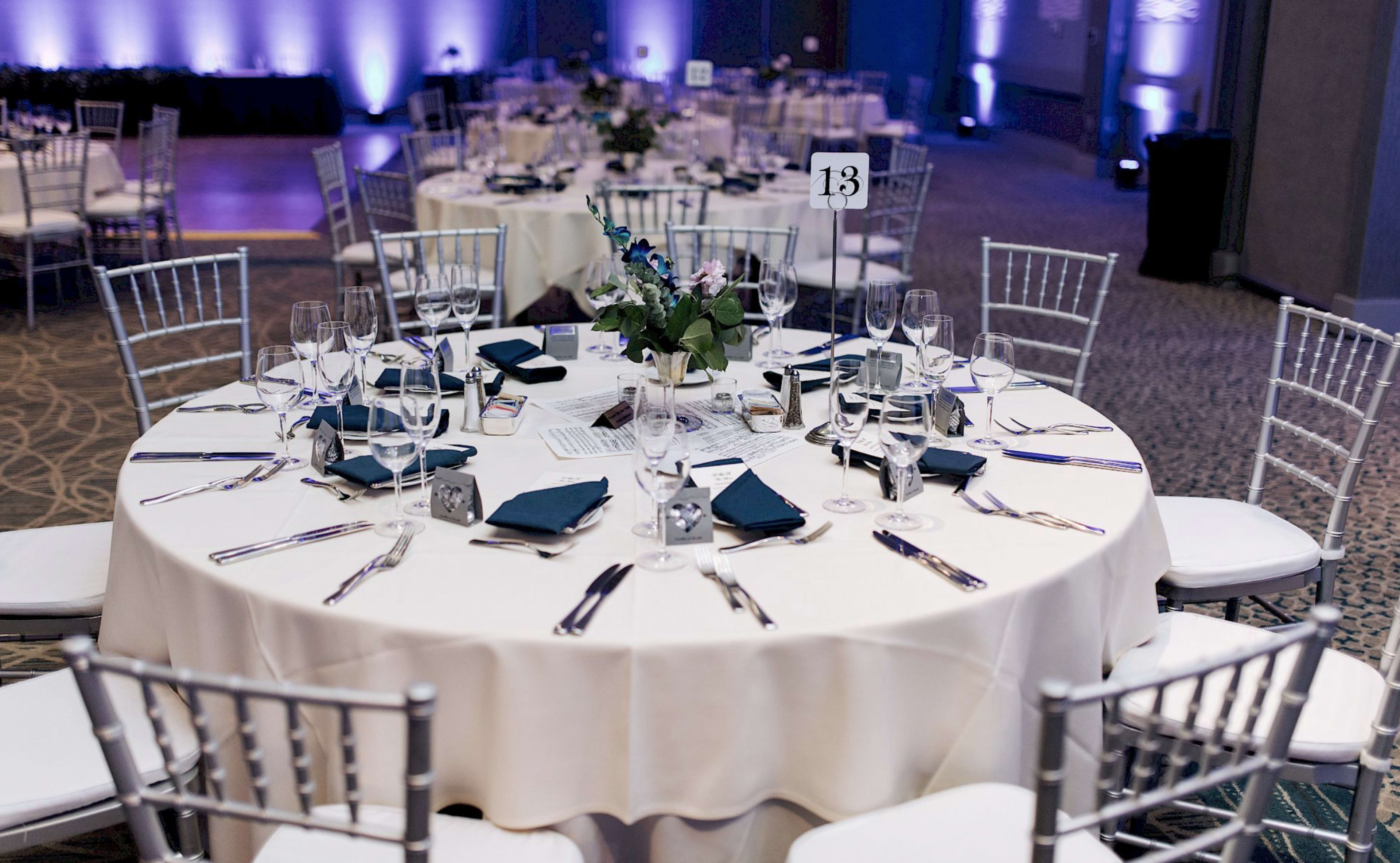 A round banquet table is set with elegant dinnerware, glassware, and a small centerpiece, amidst a decorated room for an event.