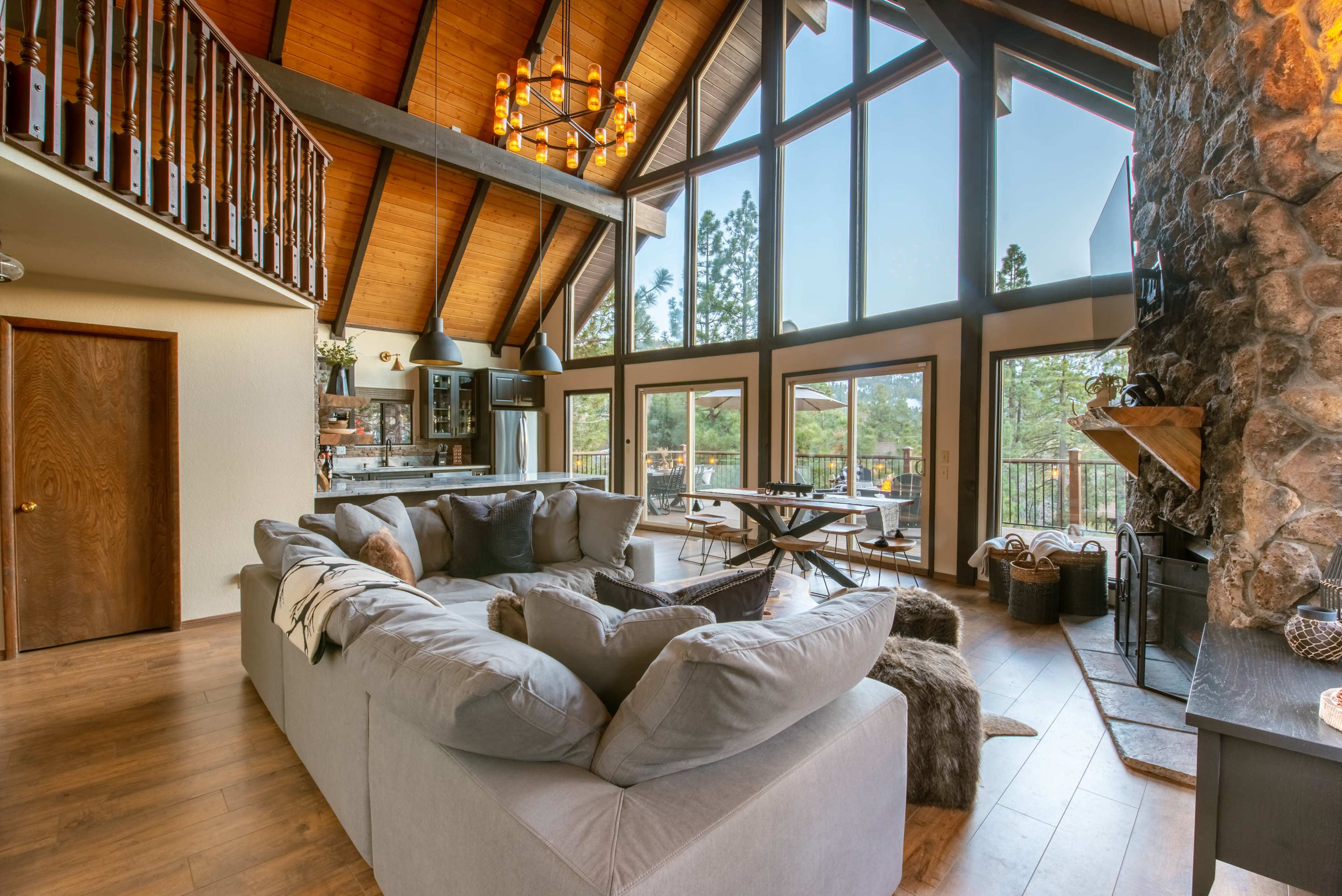 A spacious living room with a large sectional sofa, floor-to-ceiling windows, and a stone fireplace surrounded by wooden beams.