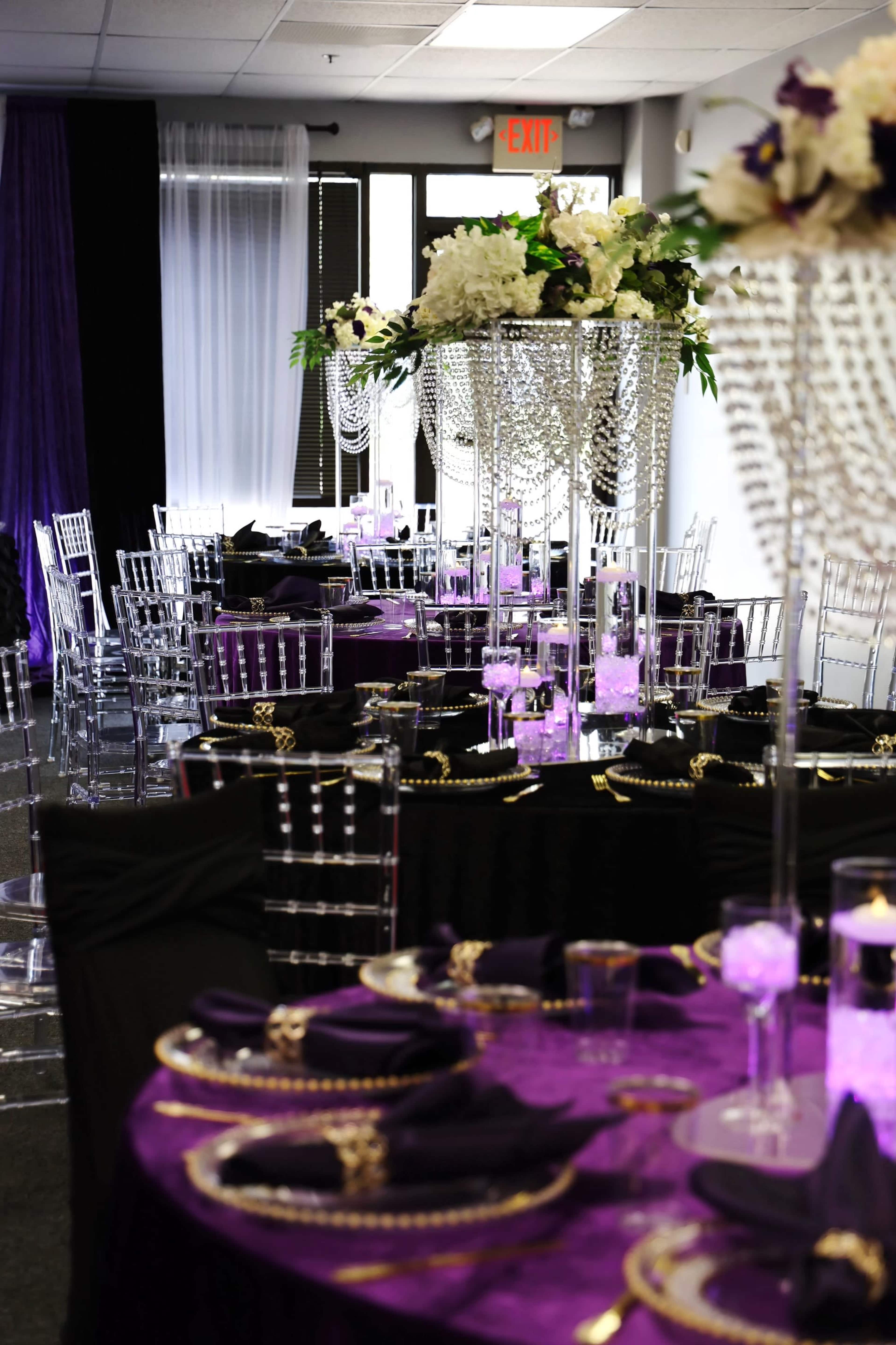 A banquet hall is set up with elegant tables covered in purple and black linens, adorned with crystal centerpieces and surrounded by clear acrylic chairs.