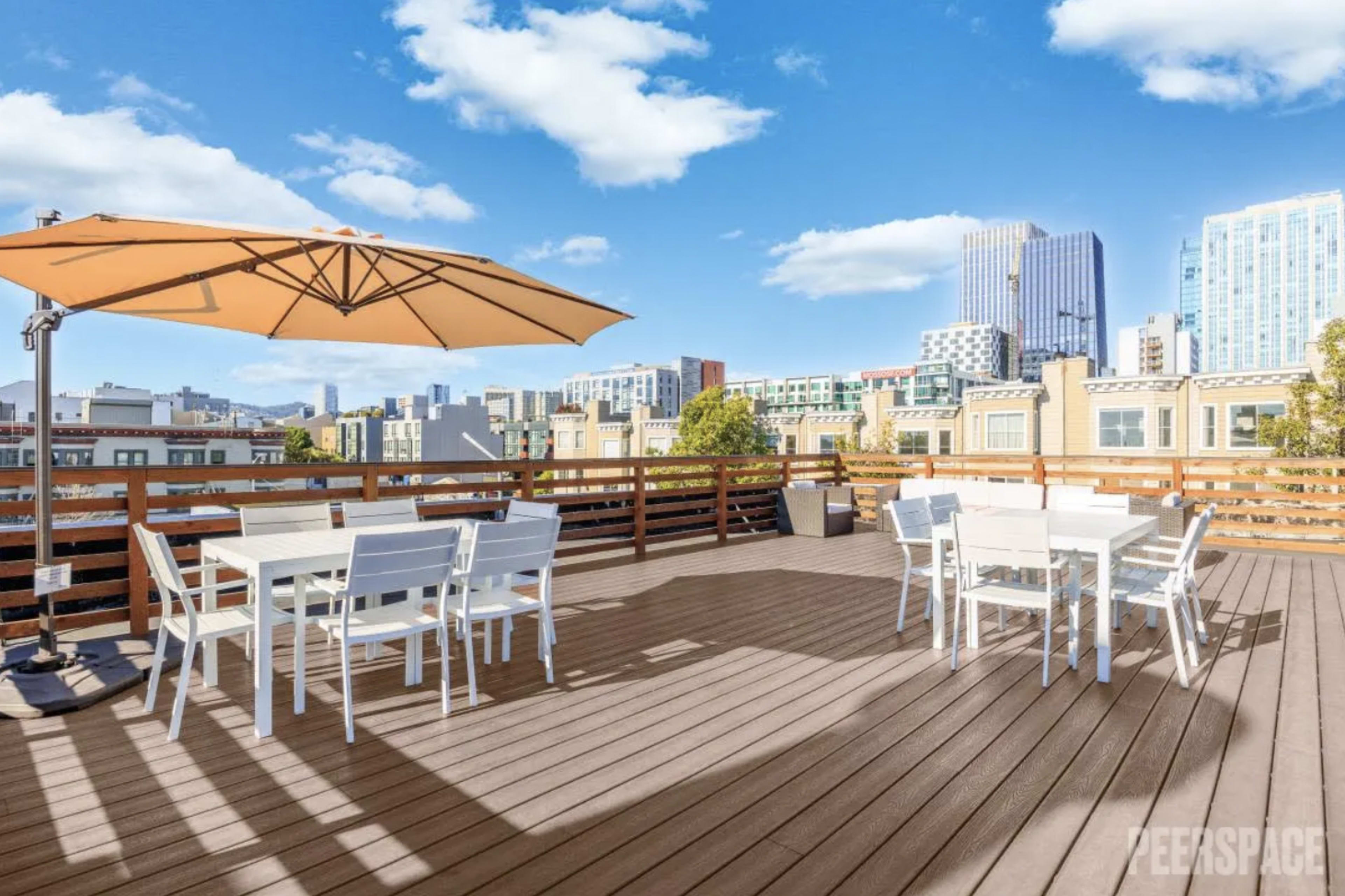 The image shows a spacious rooftop deck with white outdoor furniture and a large umbrella, overlooking a city skyline under a blue sky.