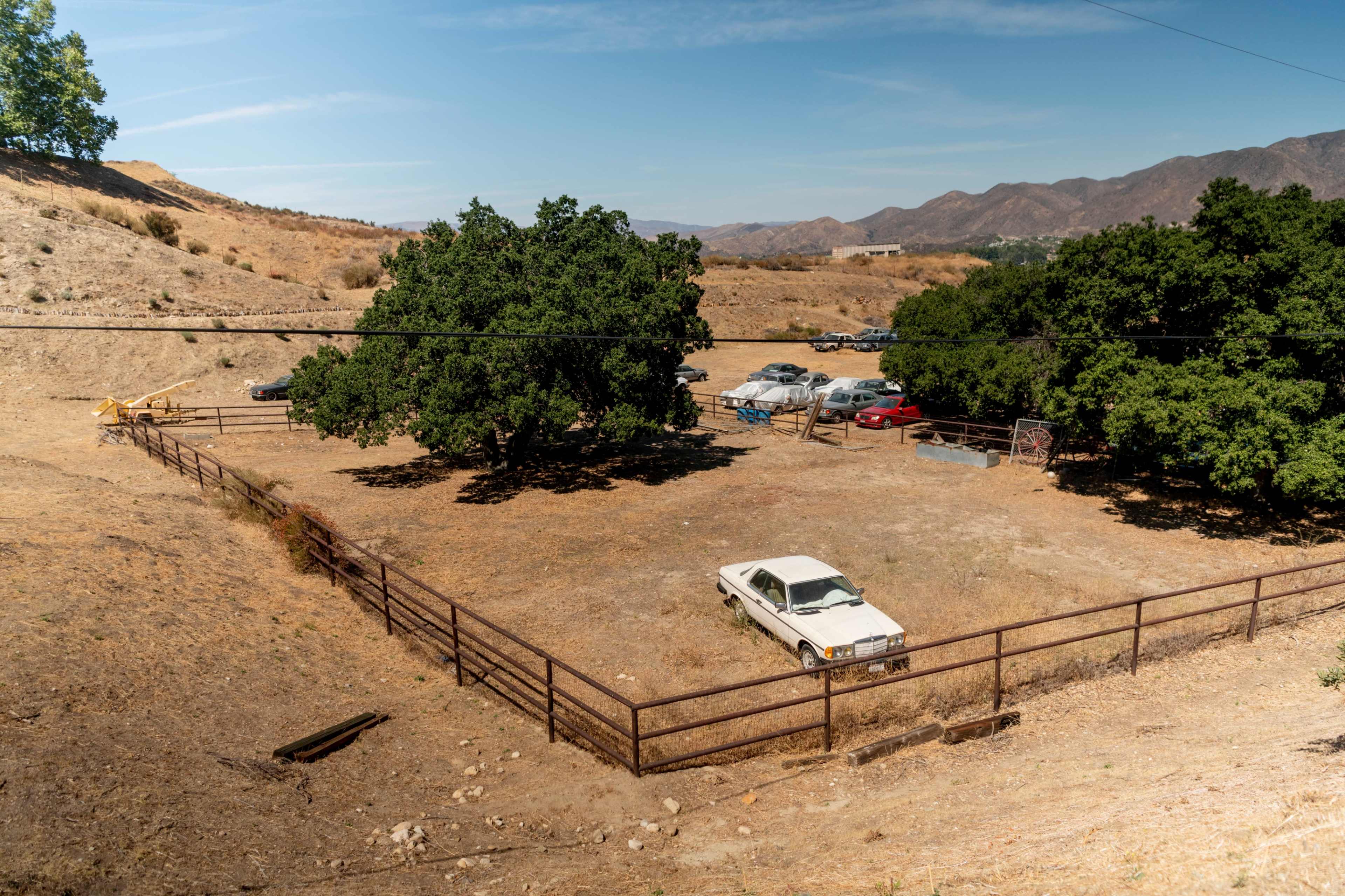 A fenced dirt lot contains an old white car surrounded by sparse vegetation and distant mountains.