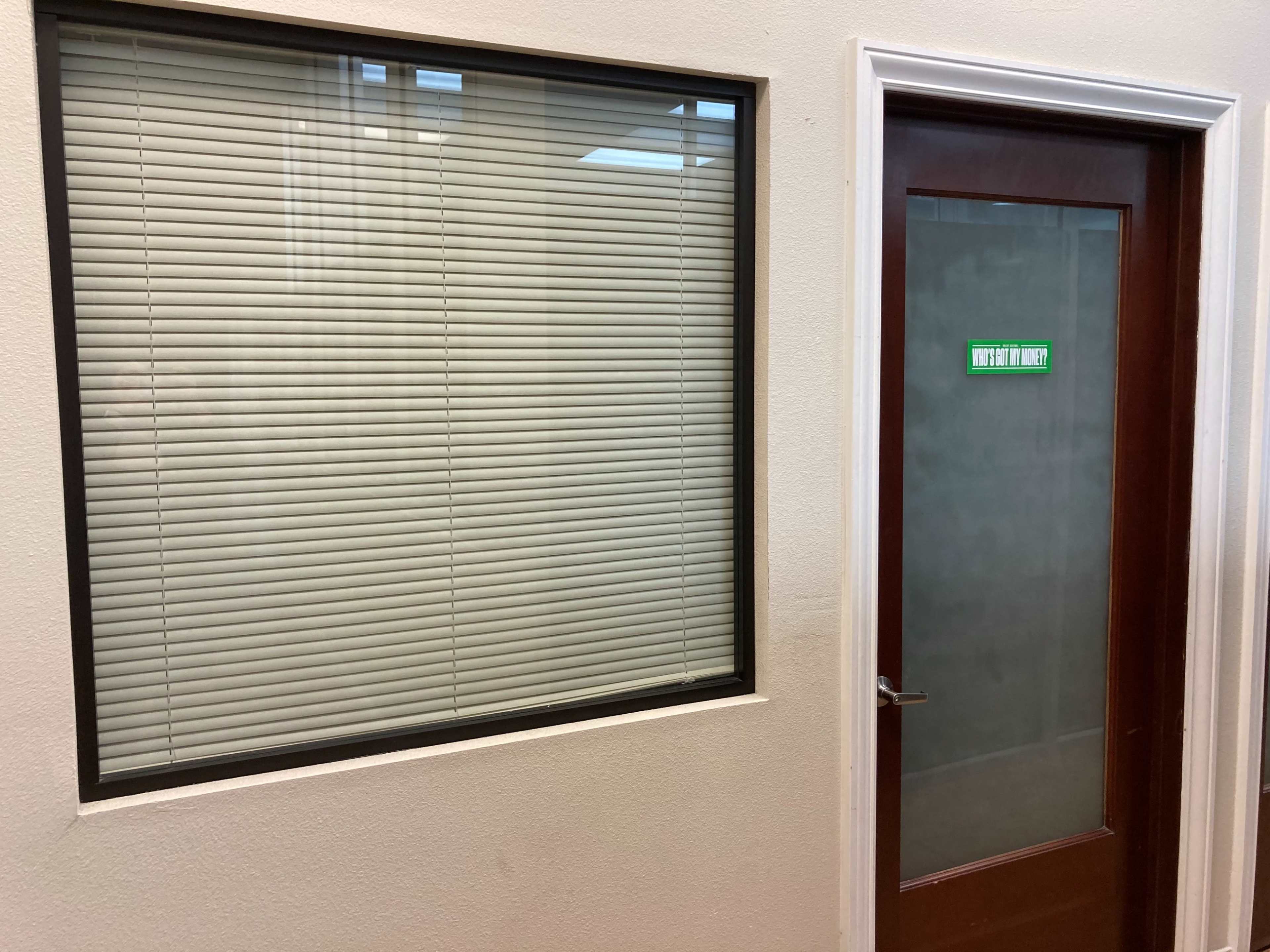 A wall with a large window covered by blinds next to a wooden door with a green sign.