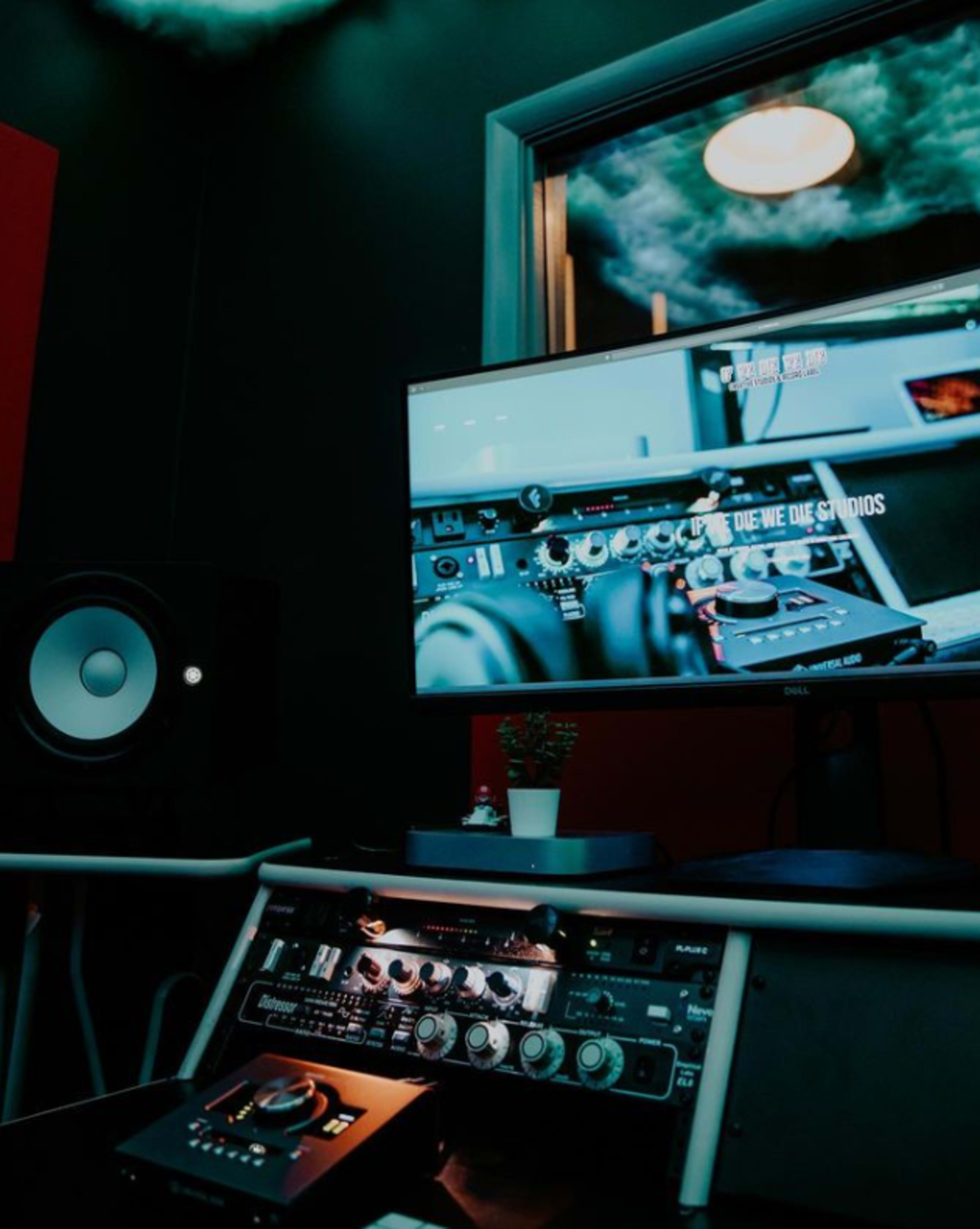 A music production setup featuring a monitor displaying audio equipment, a black audio interface with various controls, and a large speaker on a table in a dark room.