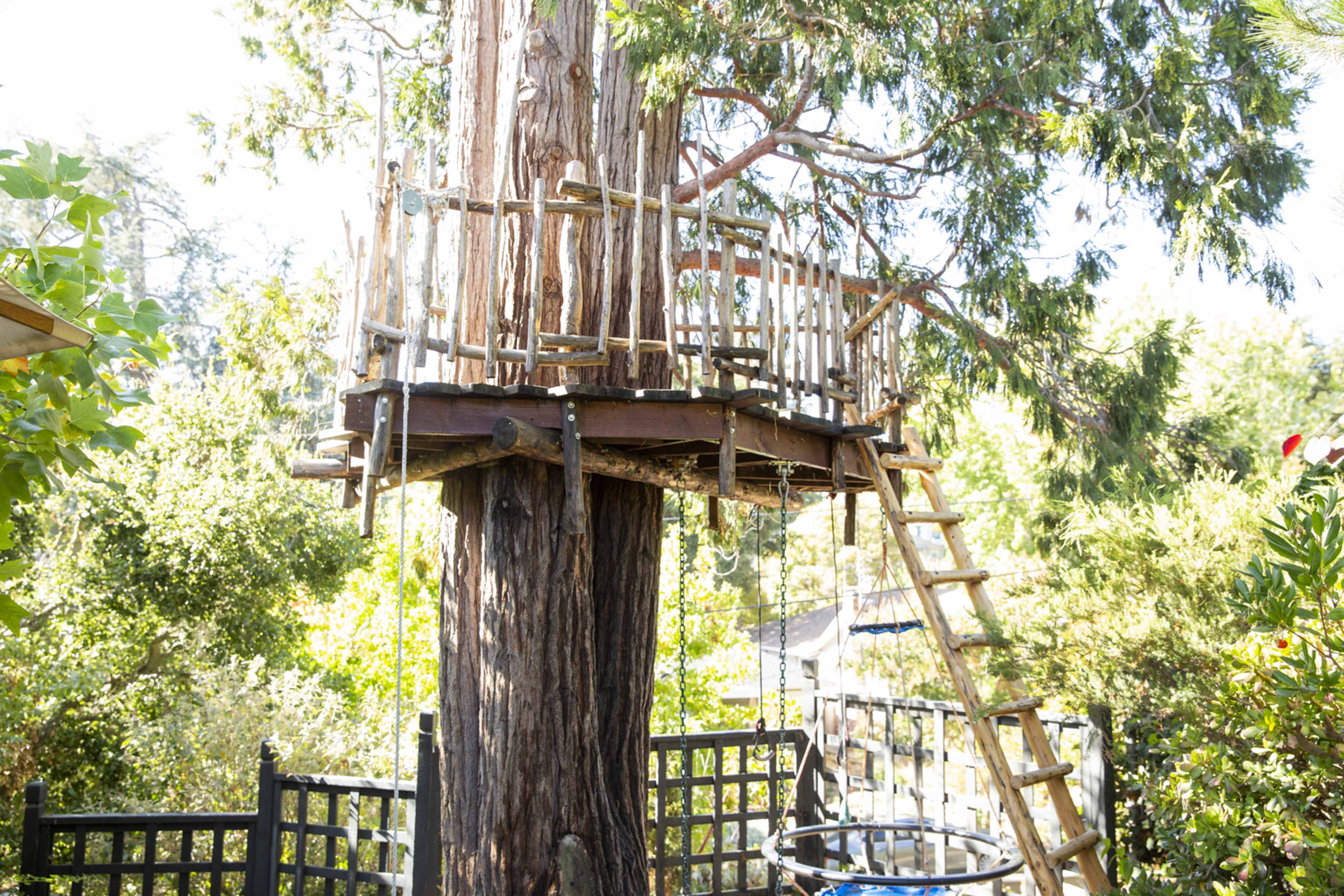 A wooden treehouse is built around a large tree, featuring a ladder and swings nearby.