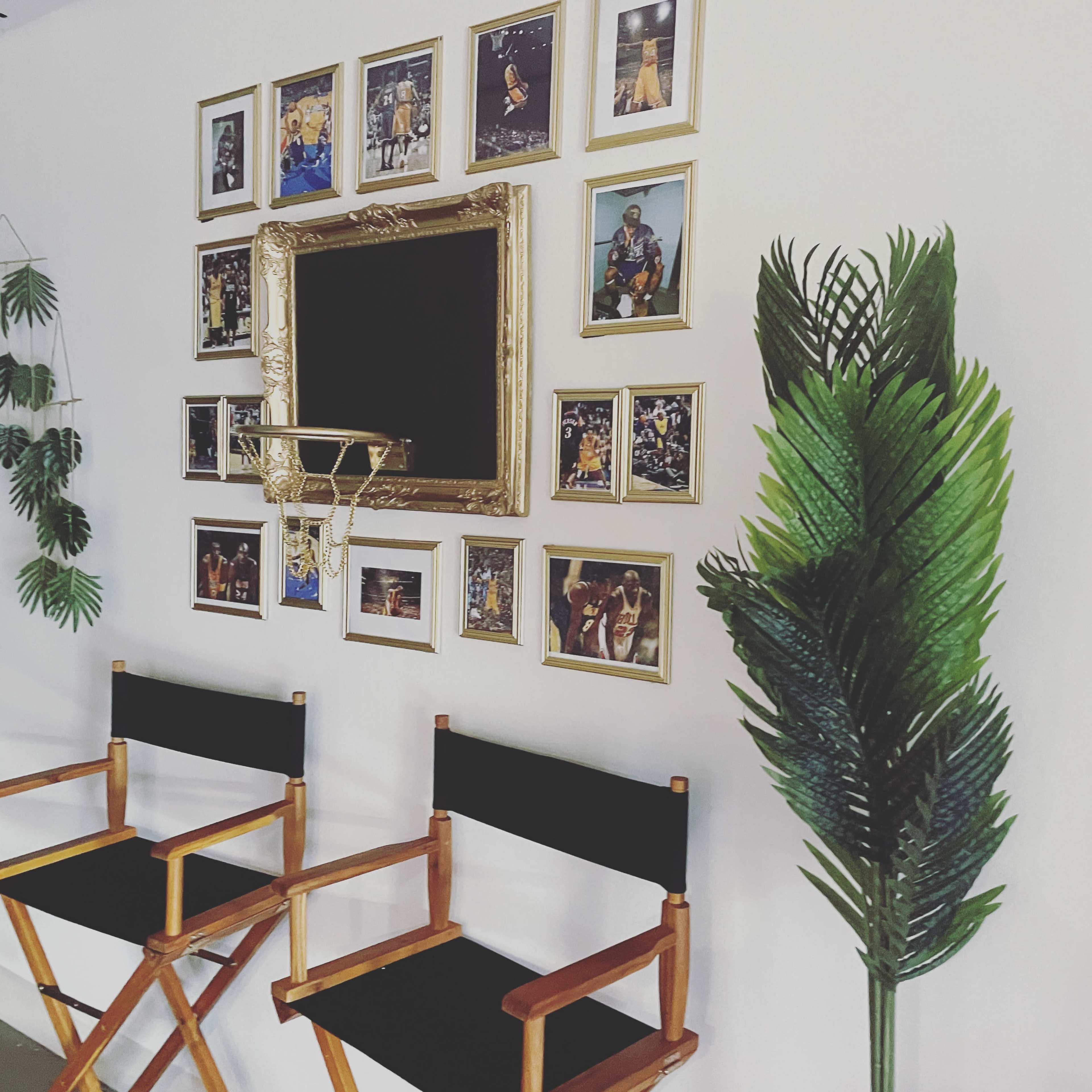 A wall adorned with multiple framed photographs, a central ornate mirror, two wooden director's chairs, and a tall green plant.