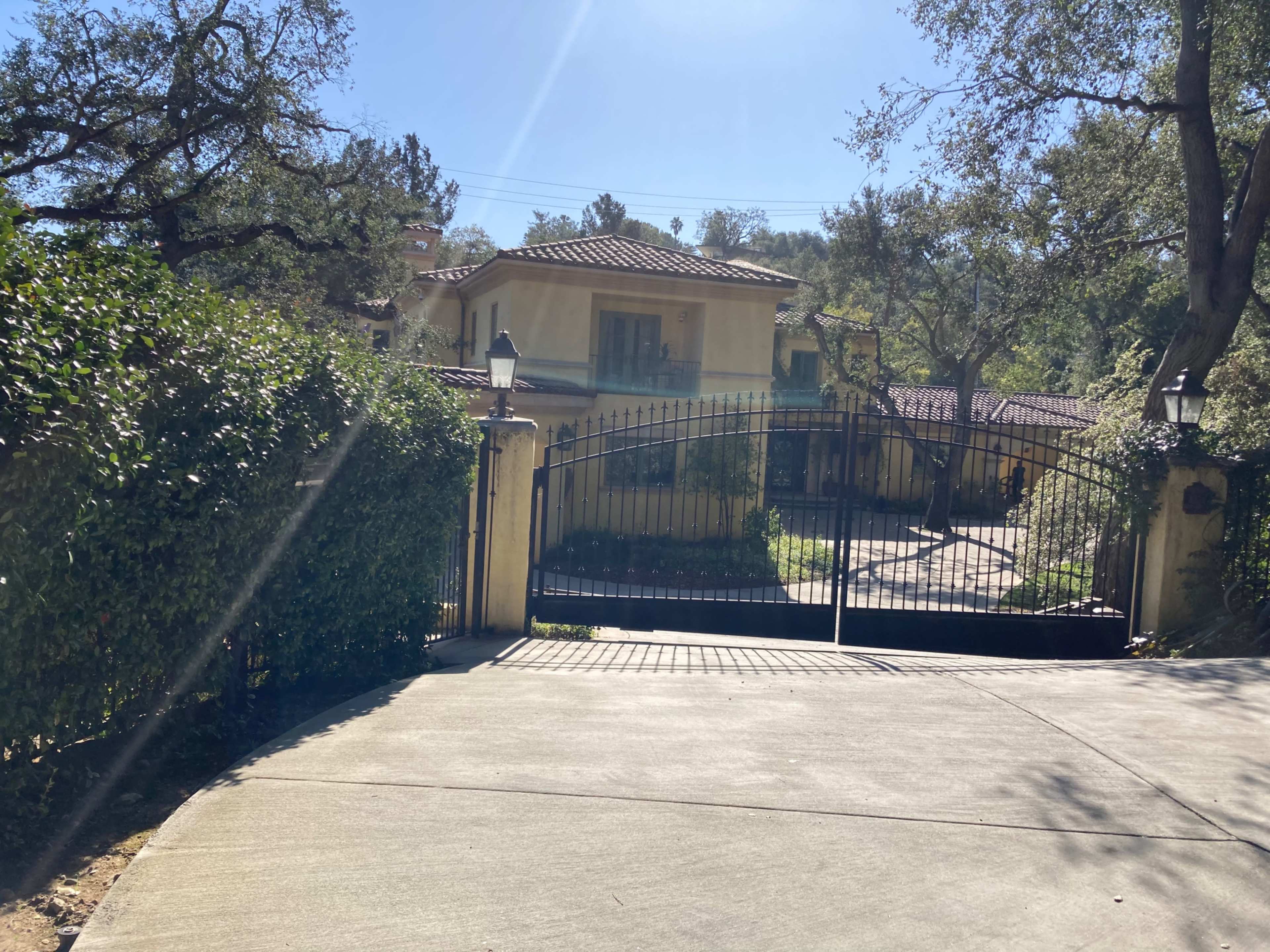 A gated driveway leads to a two-story house surrounded by trees and landscaping.