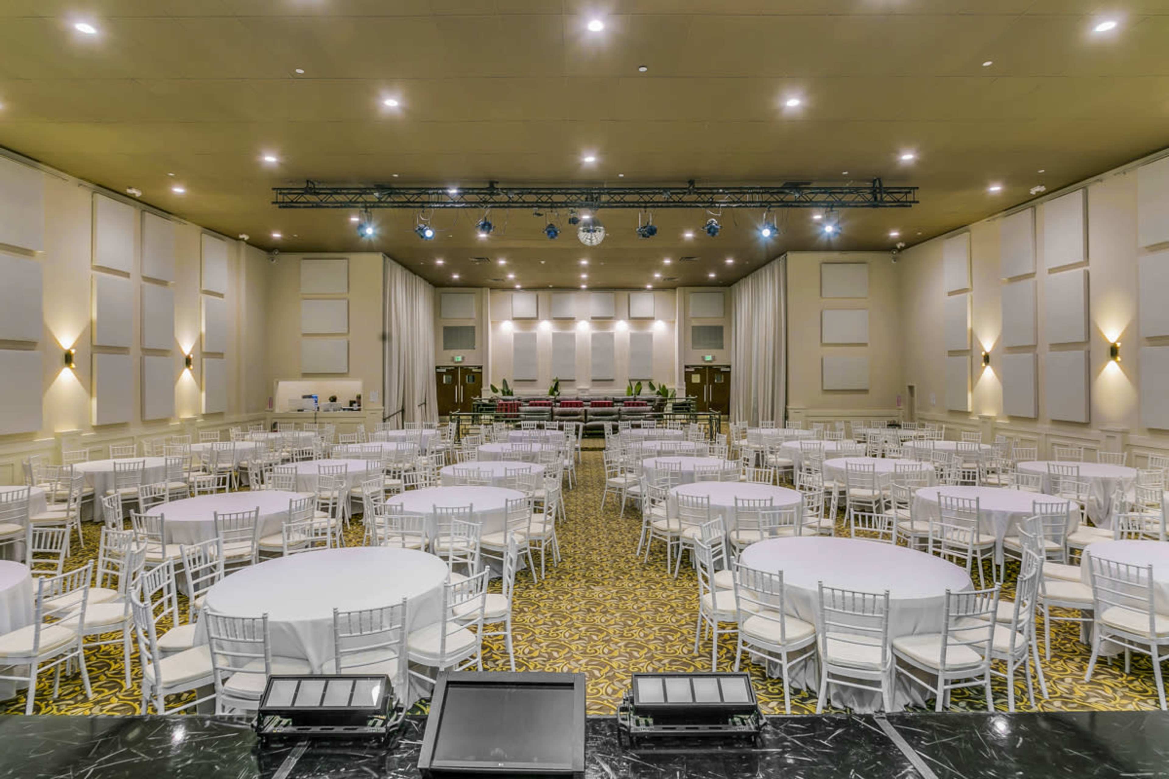 A spacious event hall filled with round tables covered in white tablecloths, surrounded by white chairs, and illuminated by ceiling lights and a disco ball.