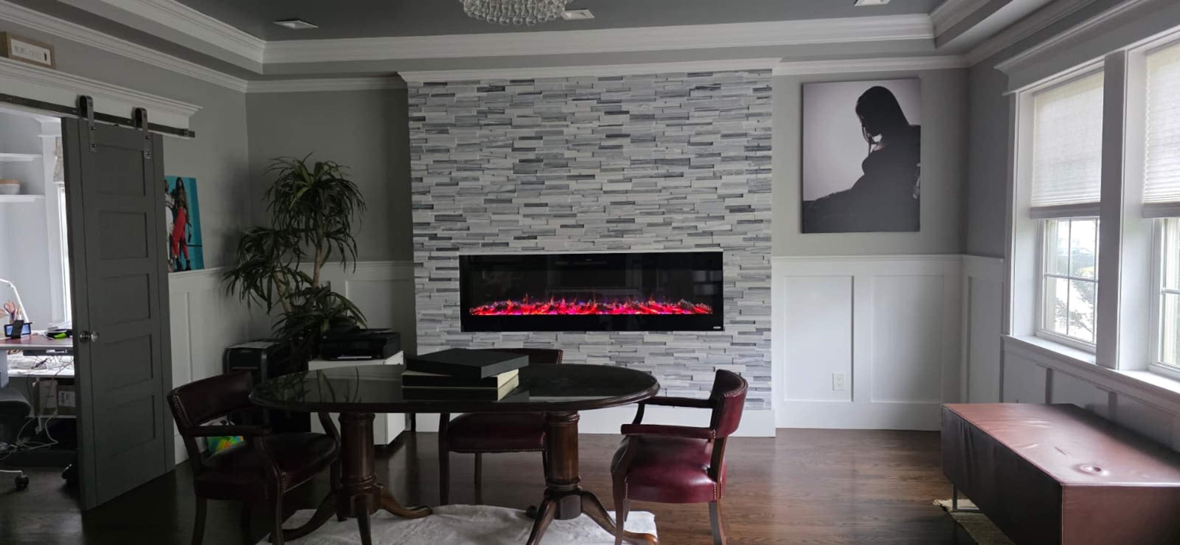 The image shows a modern living room featuring a sleek, electric fireplace with a stone facade, a round dining table surrounded by four chairs, and a large window with shades.