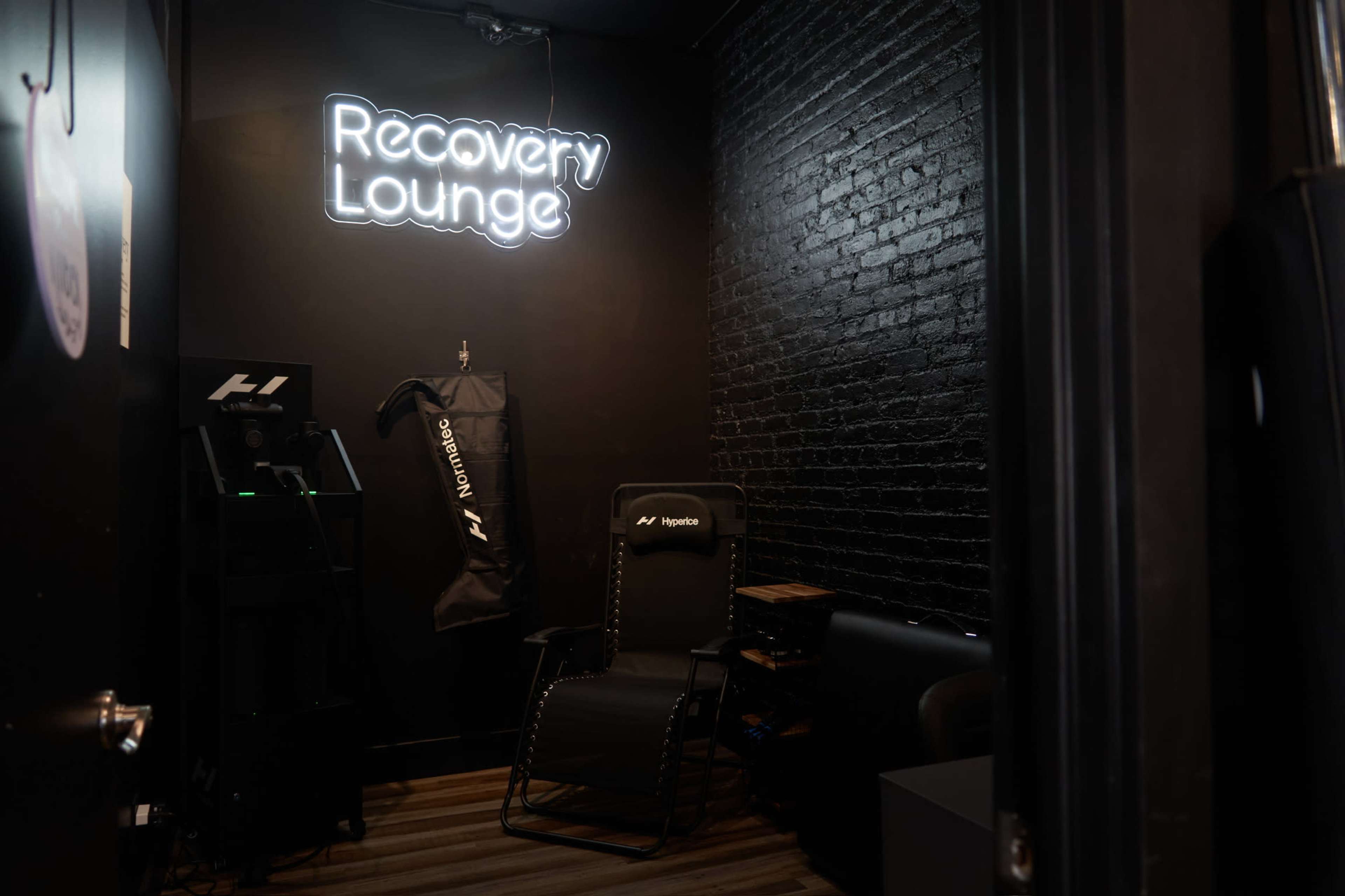 The image shows a dimly lit room with a neon sign reading "Recovery Lounge," a black brick wall, a reclining chair, and a recovery machine.