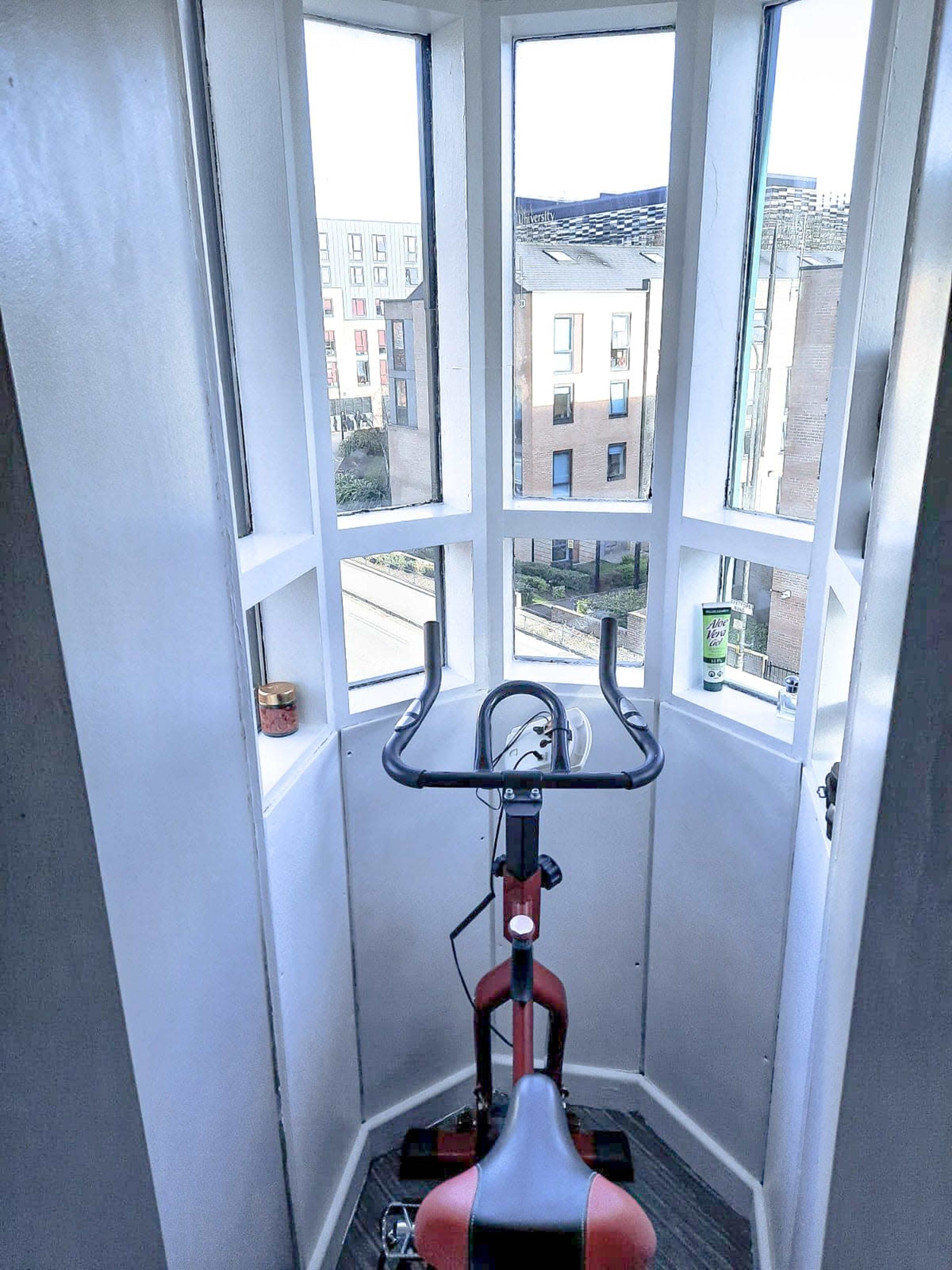 A stationary bike is positioned in a small, well-lit corner room with large windows overlooking a street and nearby buildings.