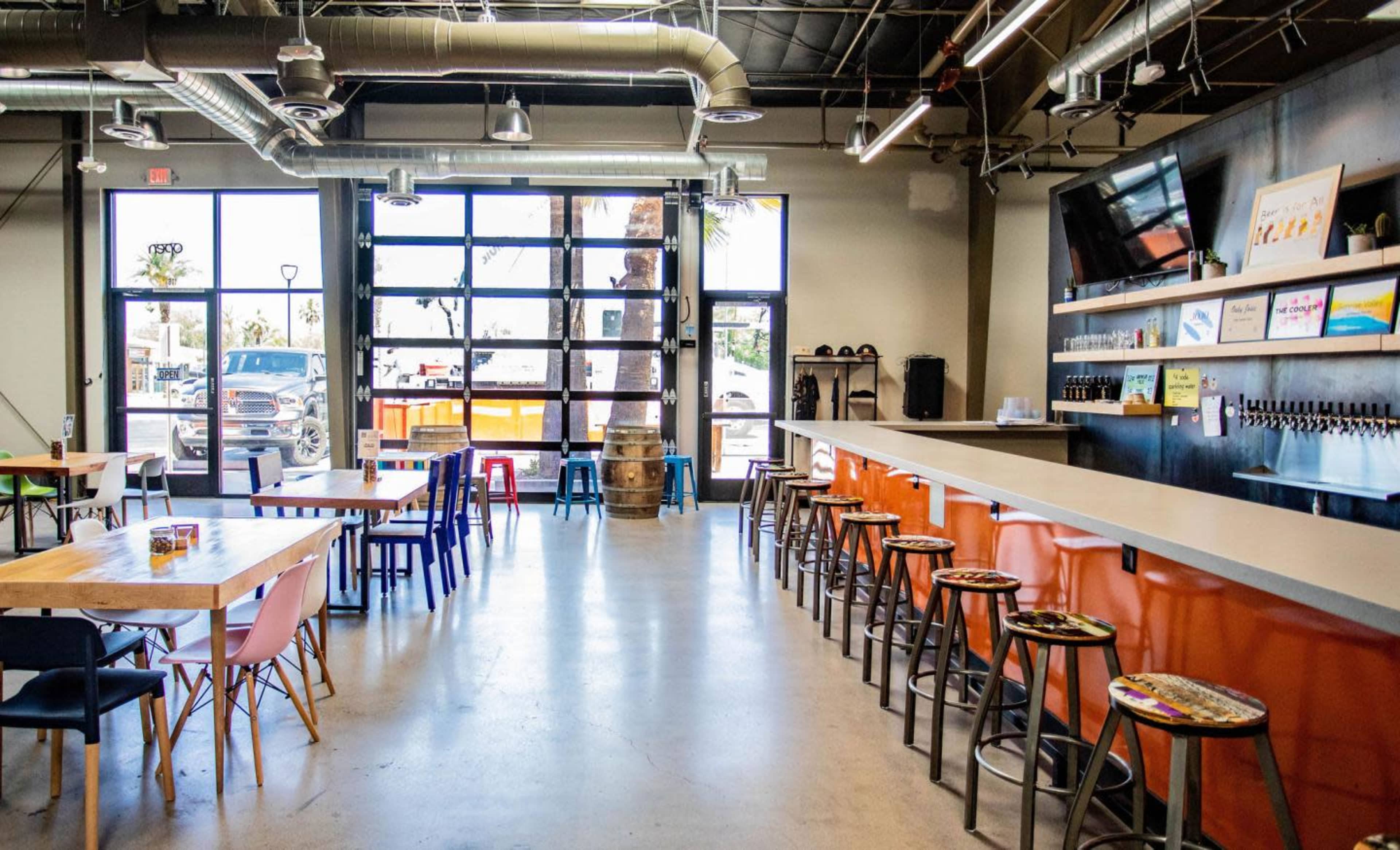 The image shows a spacious indoor seating area of a bar with modern stools and a long counter, featuring large windows that let in natural light.