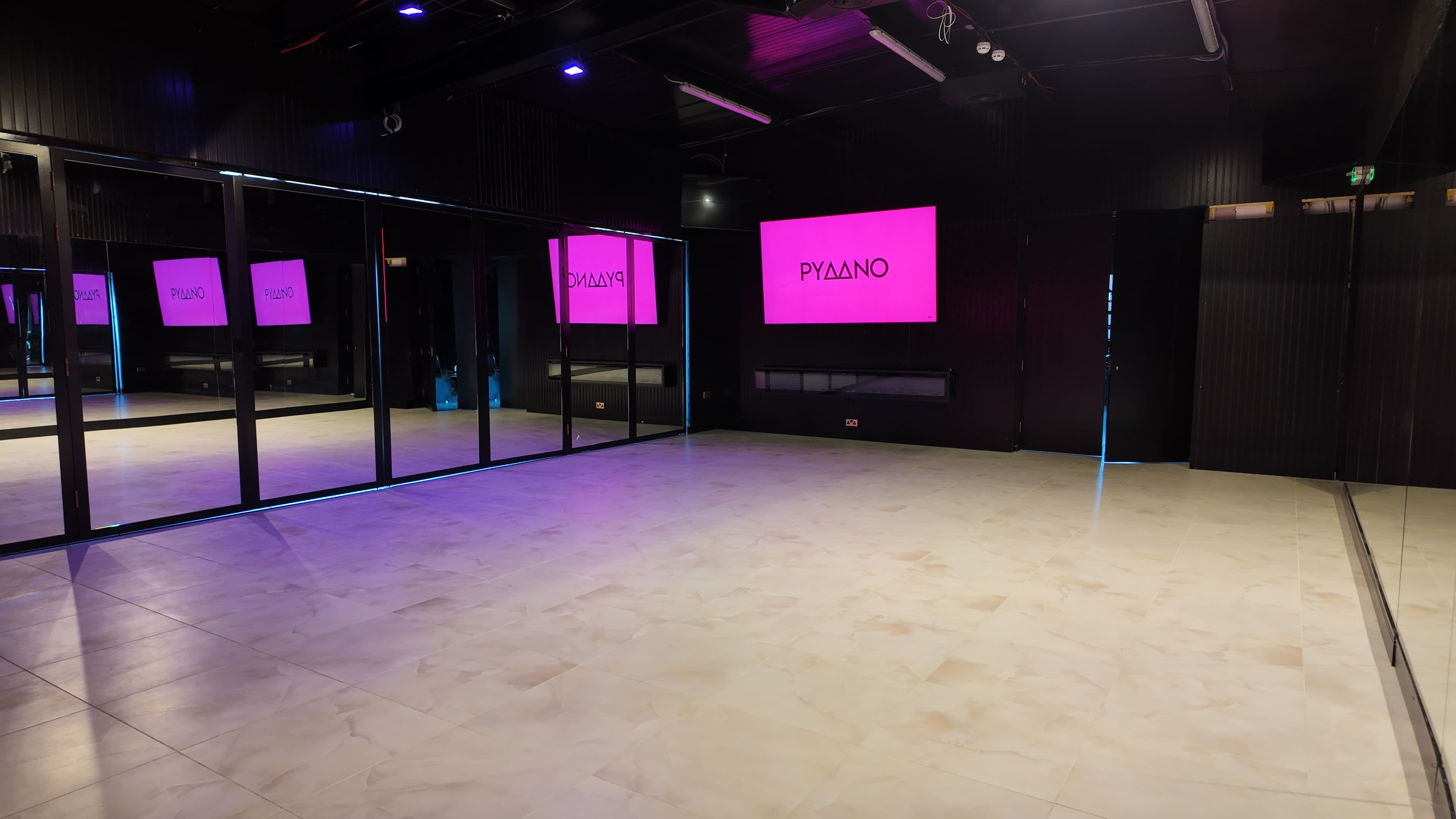 The image shows a spacious dance studio with mirrored walls, a light-colored floor, and a prominent pink illuminated sign that reads "PYZANO."