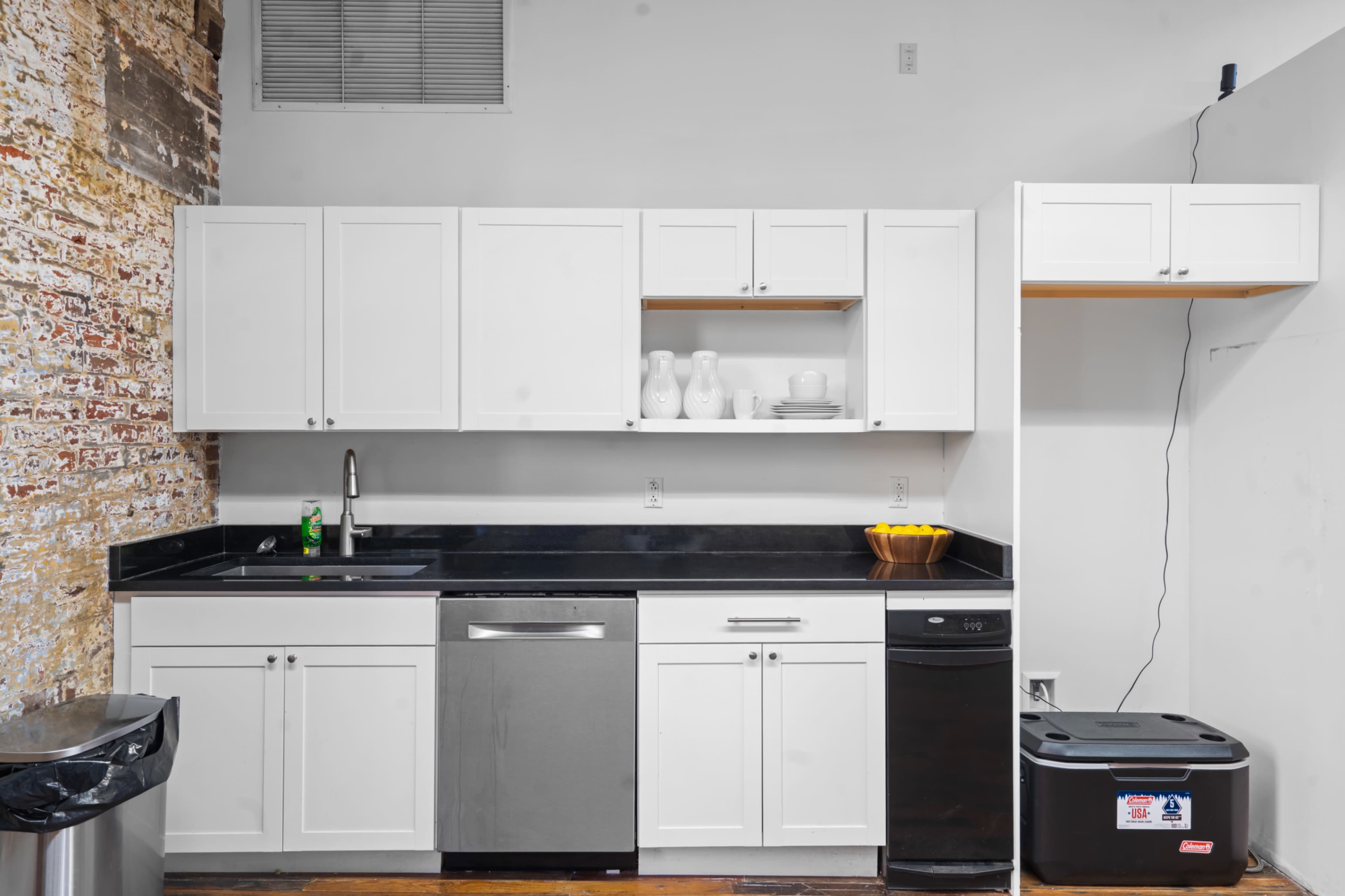 The image shows a modern kitchen with white cabinets, a black countertop, stainless steel appliances, and exposed brick walls.