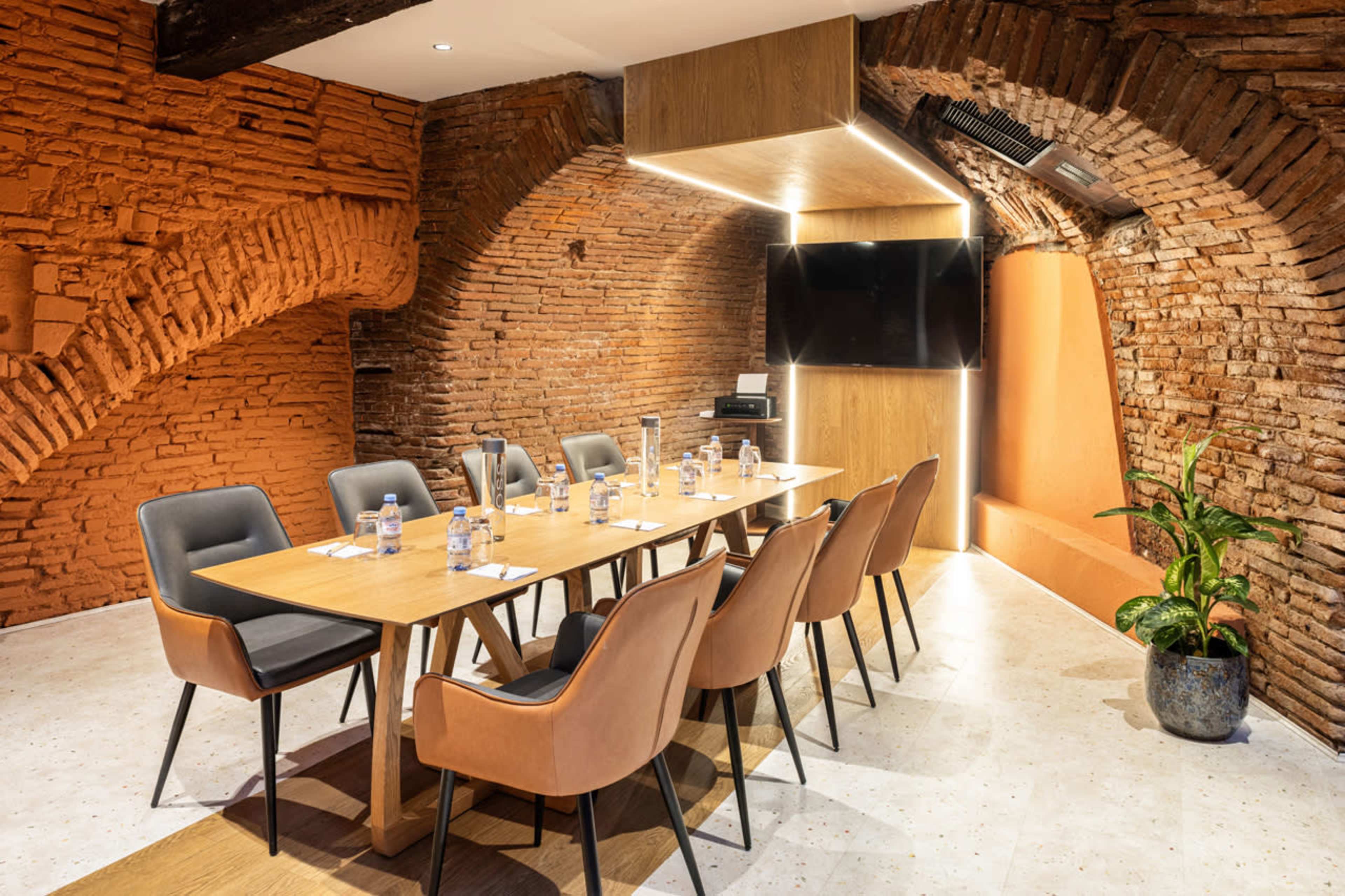 A modern conference room features a long wooden table surrounded by upholstered chairs, with exposed brick walls and a large screen mounted for presentations.