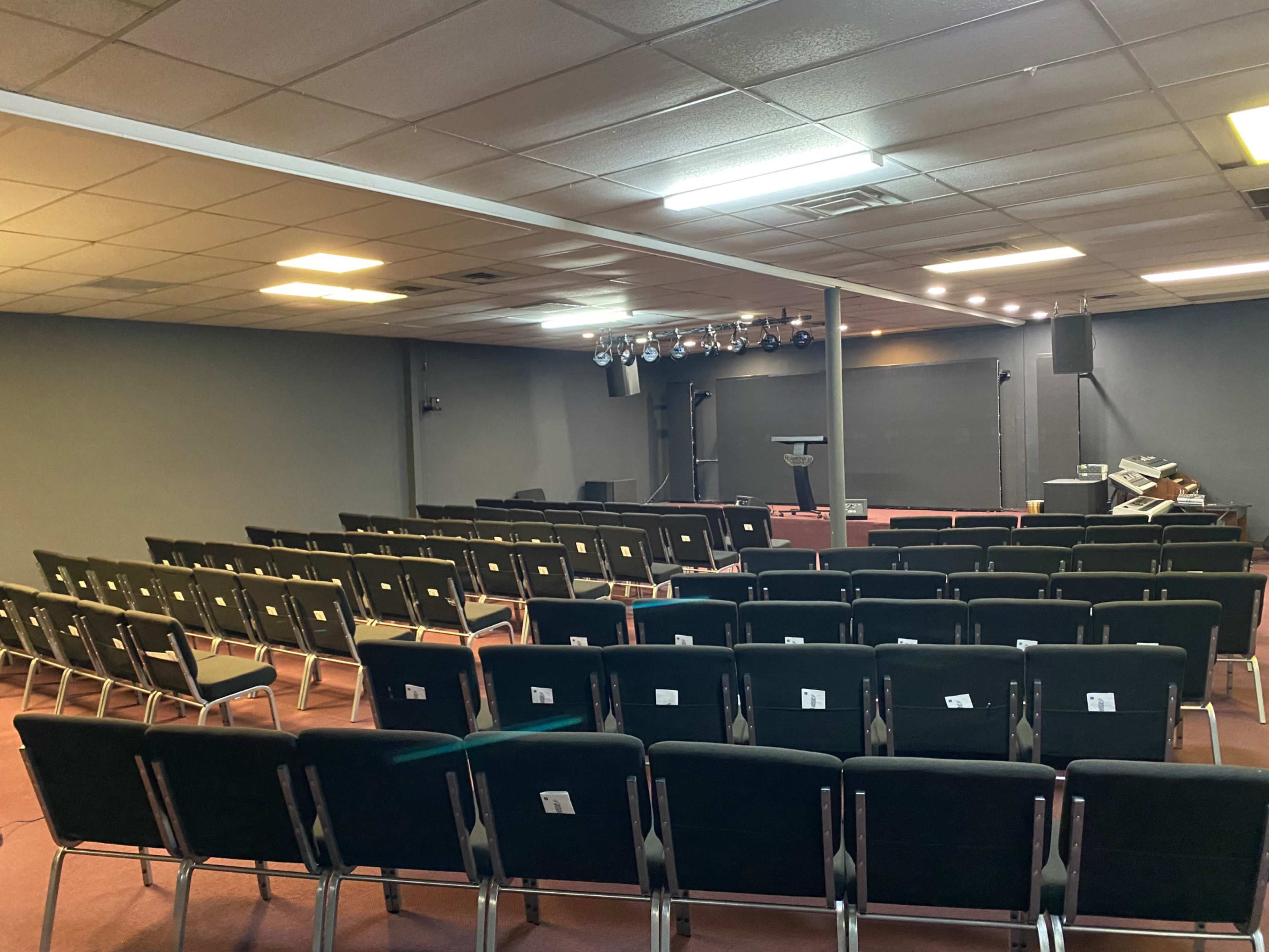 The image shows an empty auditorium with rows of black chairs facing a stage equipped with a podium and lighting equipment.