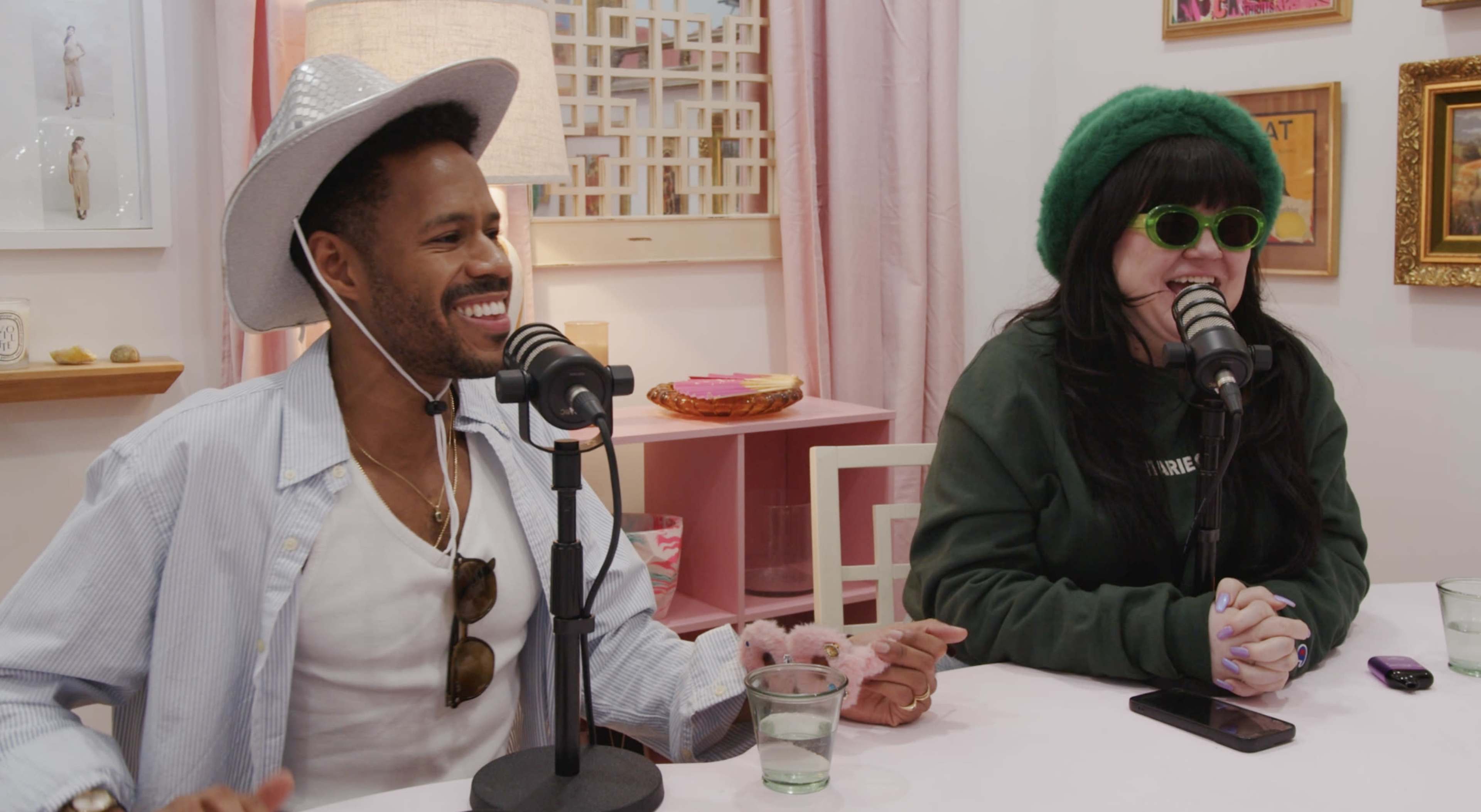 A man wearing a cowboy hat and sunglasses sits next to a woman in a green beanie and sunglasses, both speaking into microphones in a brightly decorated room.