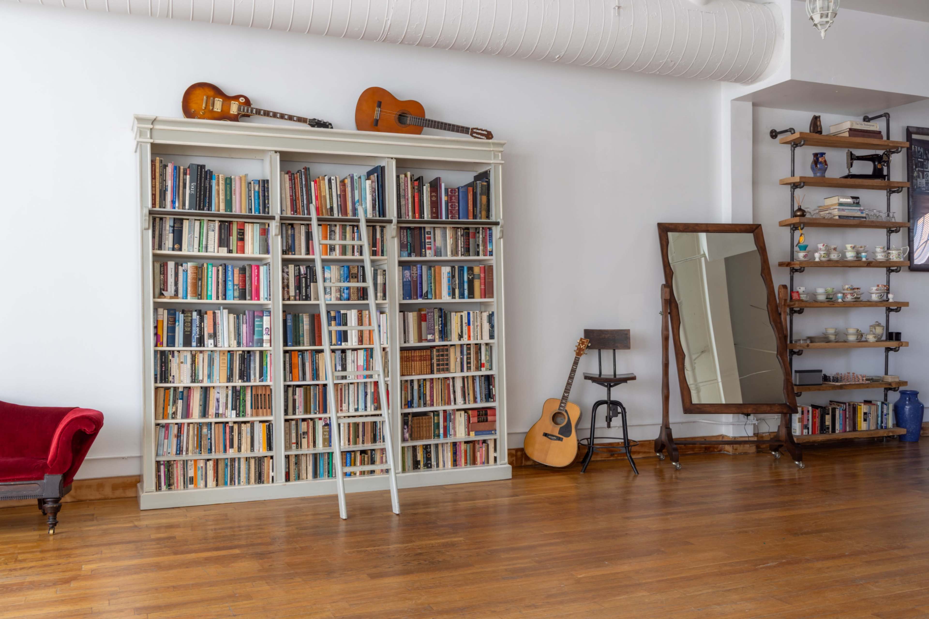A large bookshelf filled with books stands against a wall, accompanied by two guitars and a leaning ladder, while a mirror and a small table with a chair are positioned nearby.