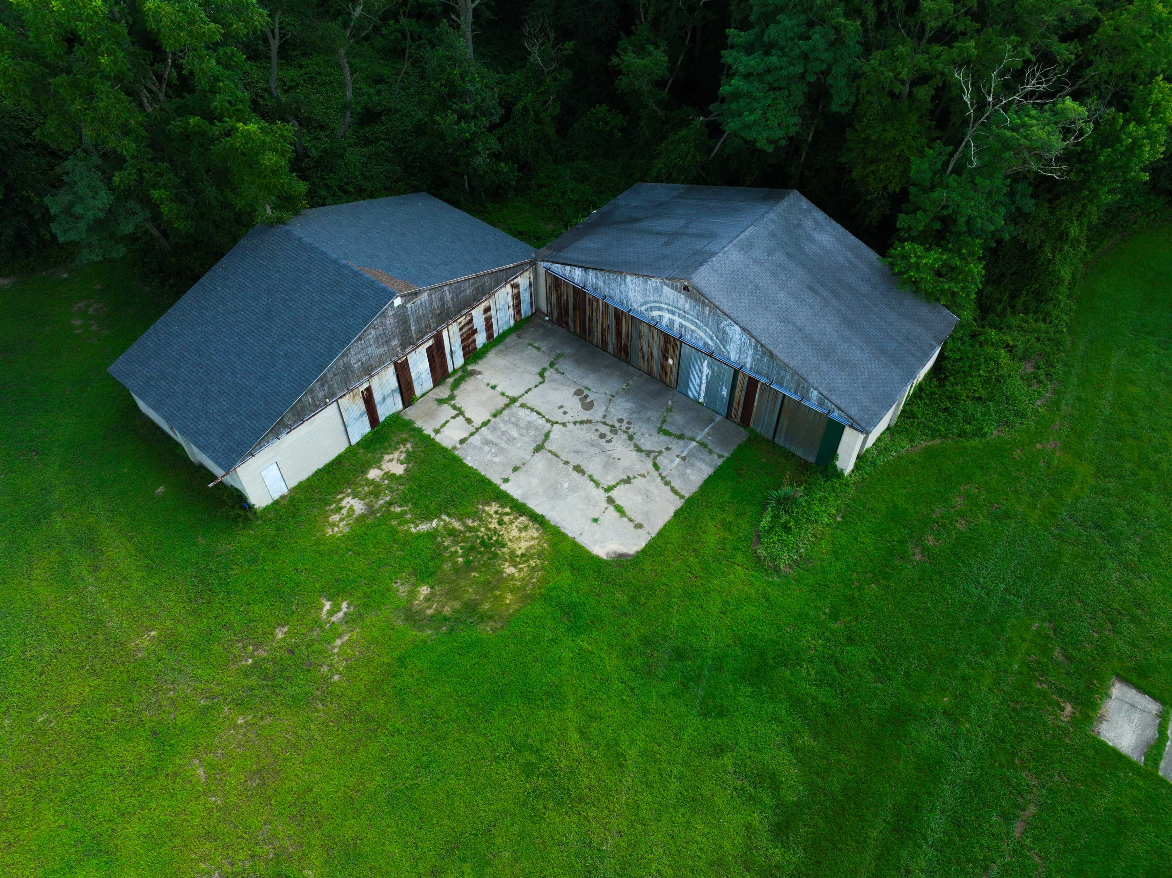The image shows two large, dilapidated buildings with boarded doors and a concrete slab, surrounded by lush green grass and trees.