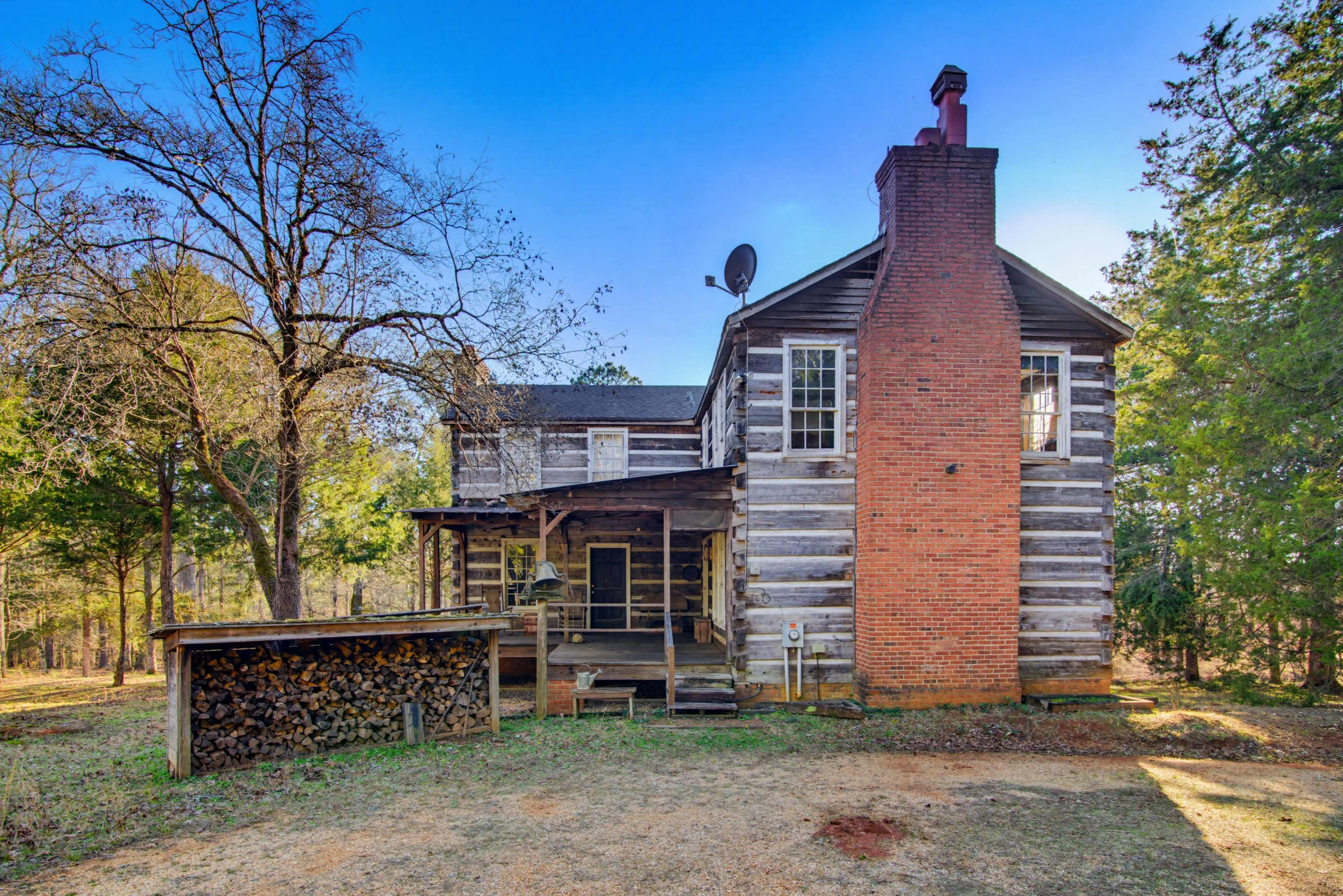 The image shows a rustic log cabin with a brick chimney, surrounded by trees, and featuring a woodpile beside the porch.