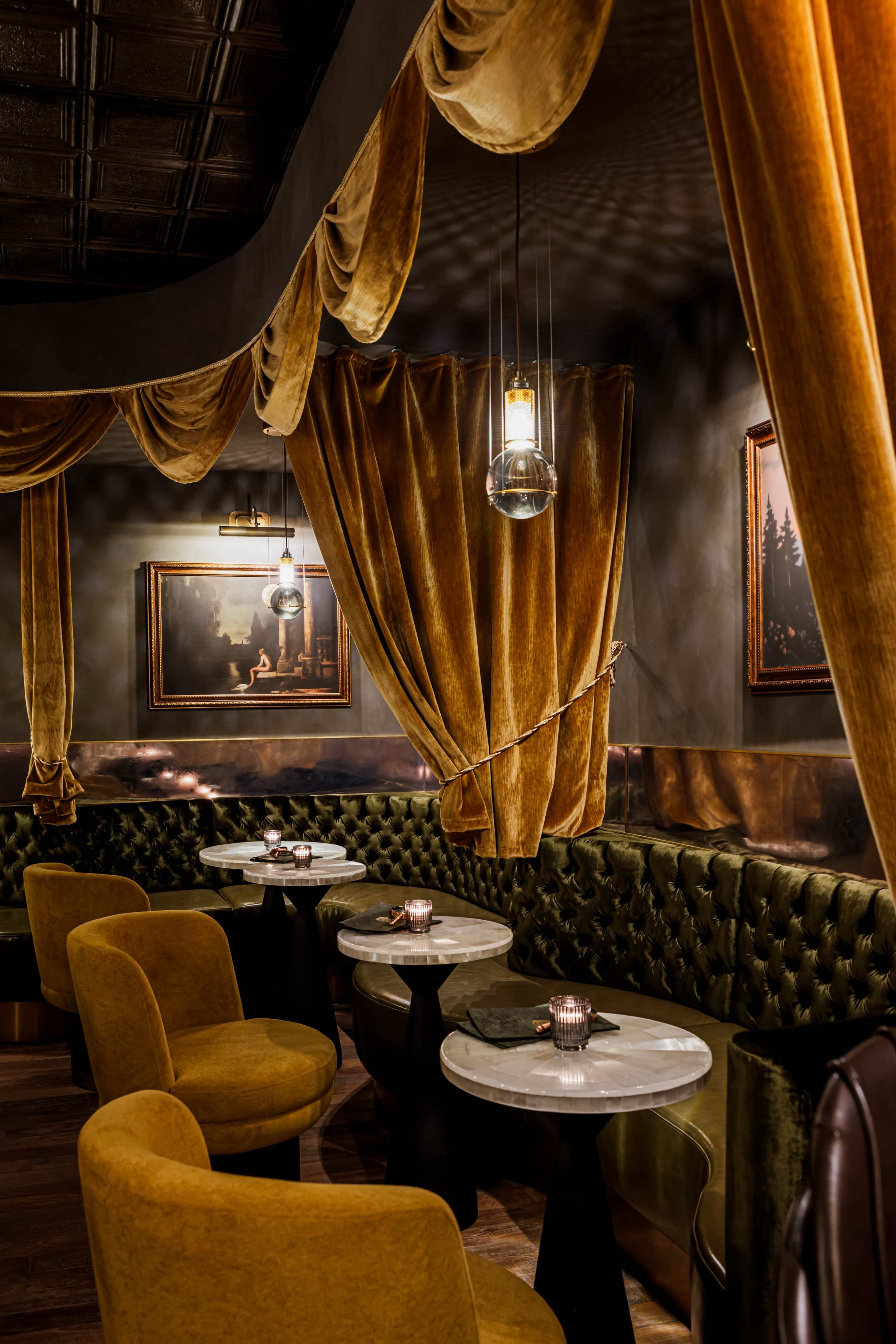 The image shows a dimly lit restaurant interior featuring green velvet seating, round marble-topped tables, and plush golden curtains.