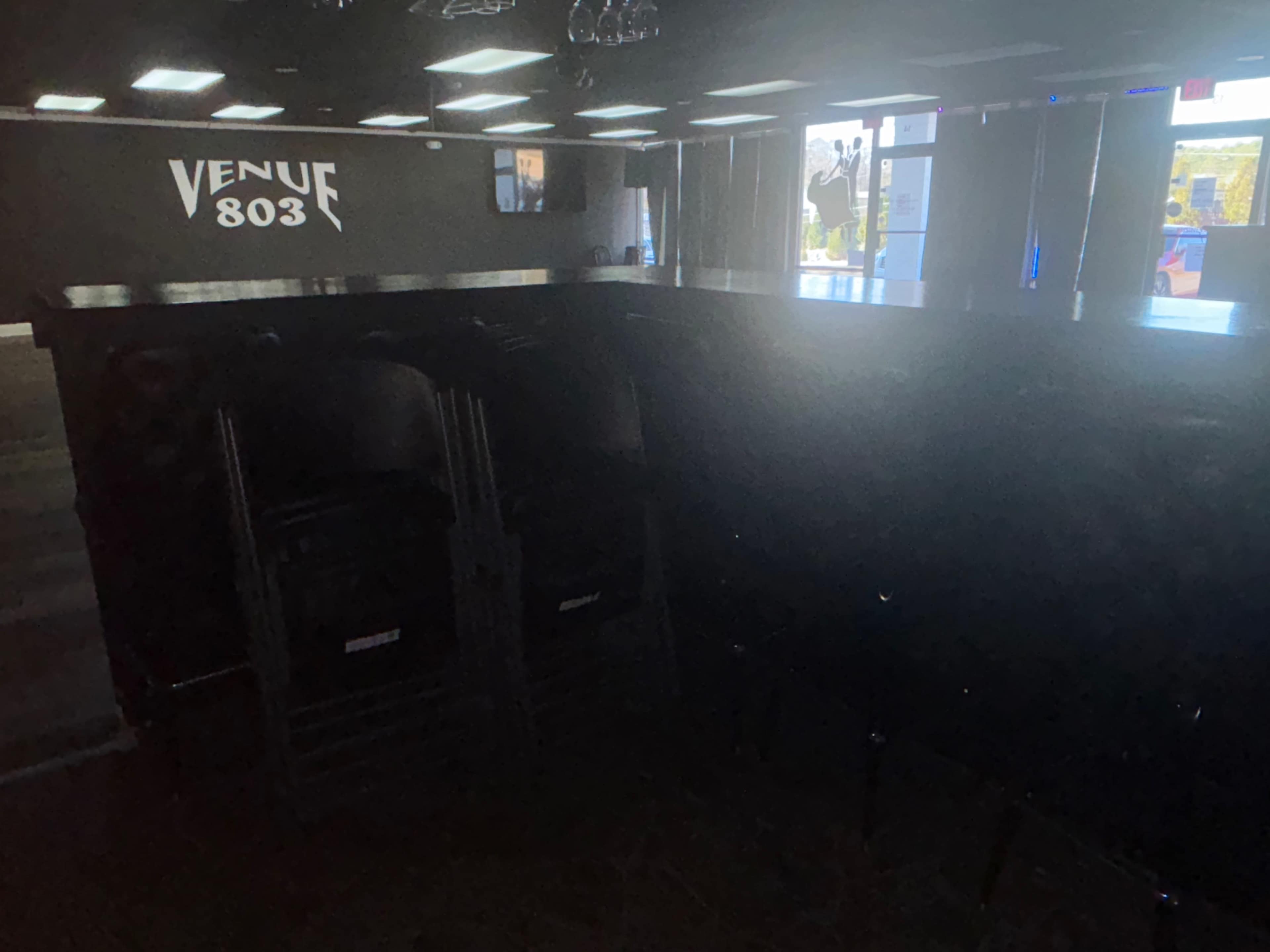 The image shows a dimly lit venue with a bar area and stacked chairs along one side.