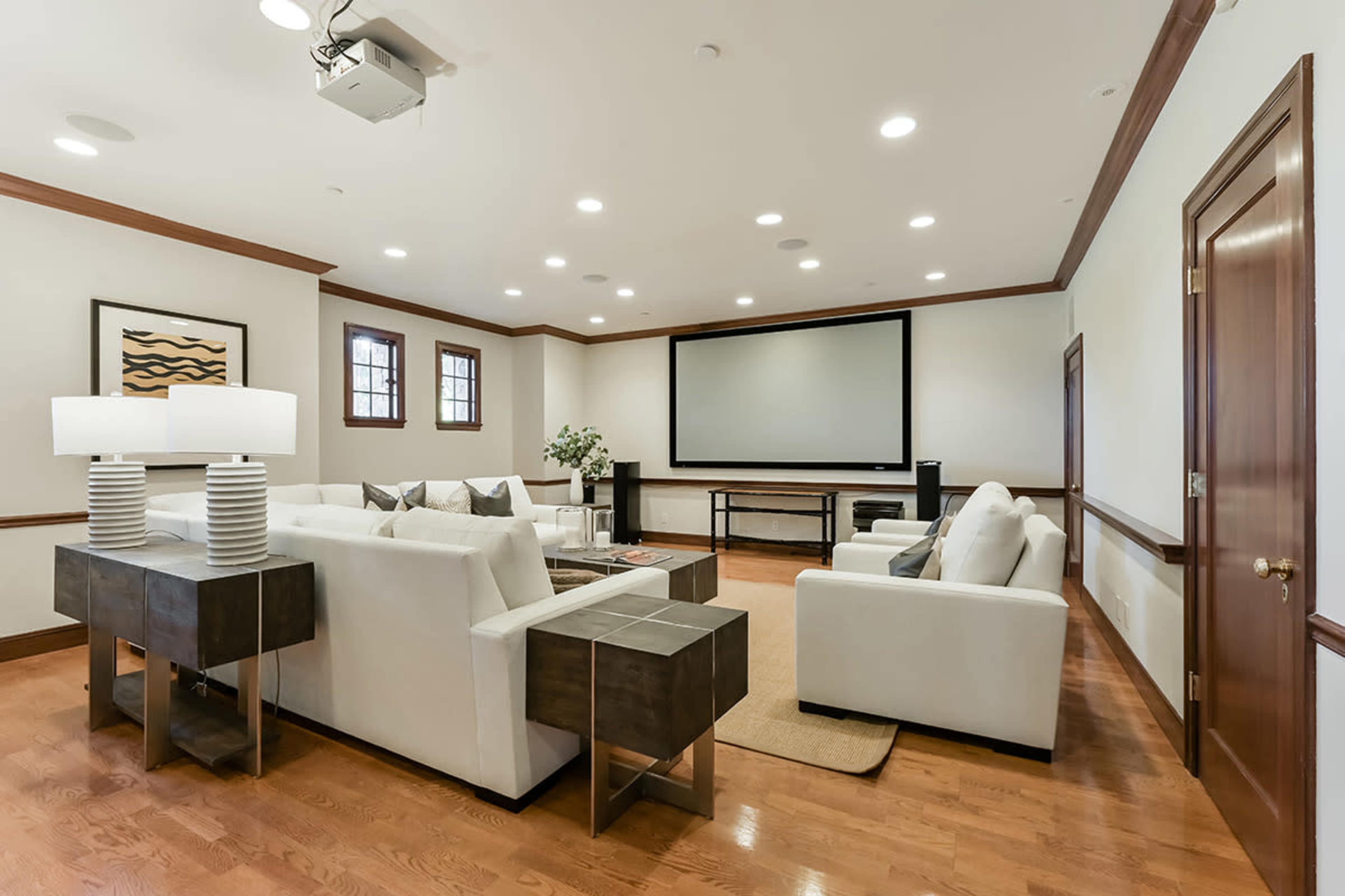 The image shows a modern living room with white sofas, a large projection screen, and wooden floors.