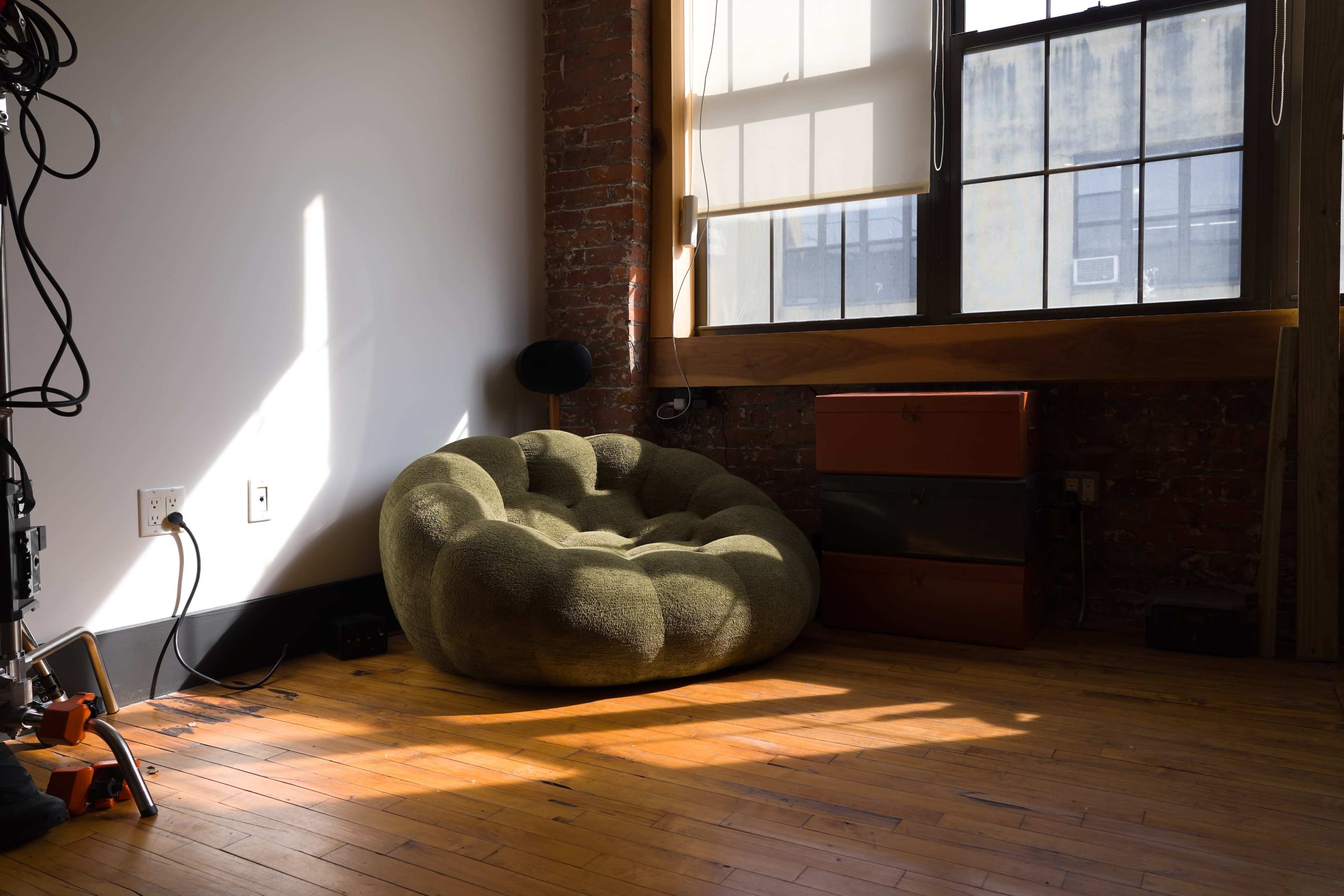 A large, round green chair is positioned in a sunlit corner of a room with wooden floors and exposed brick walls.