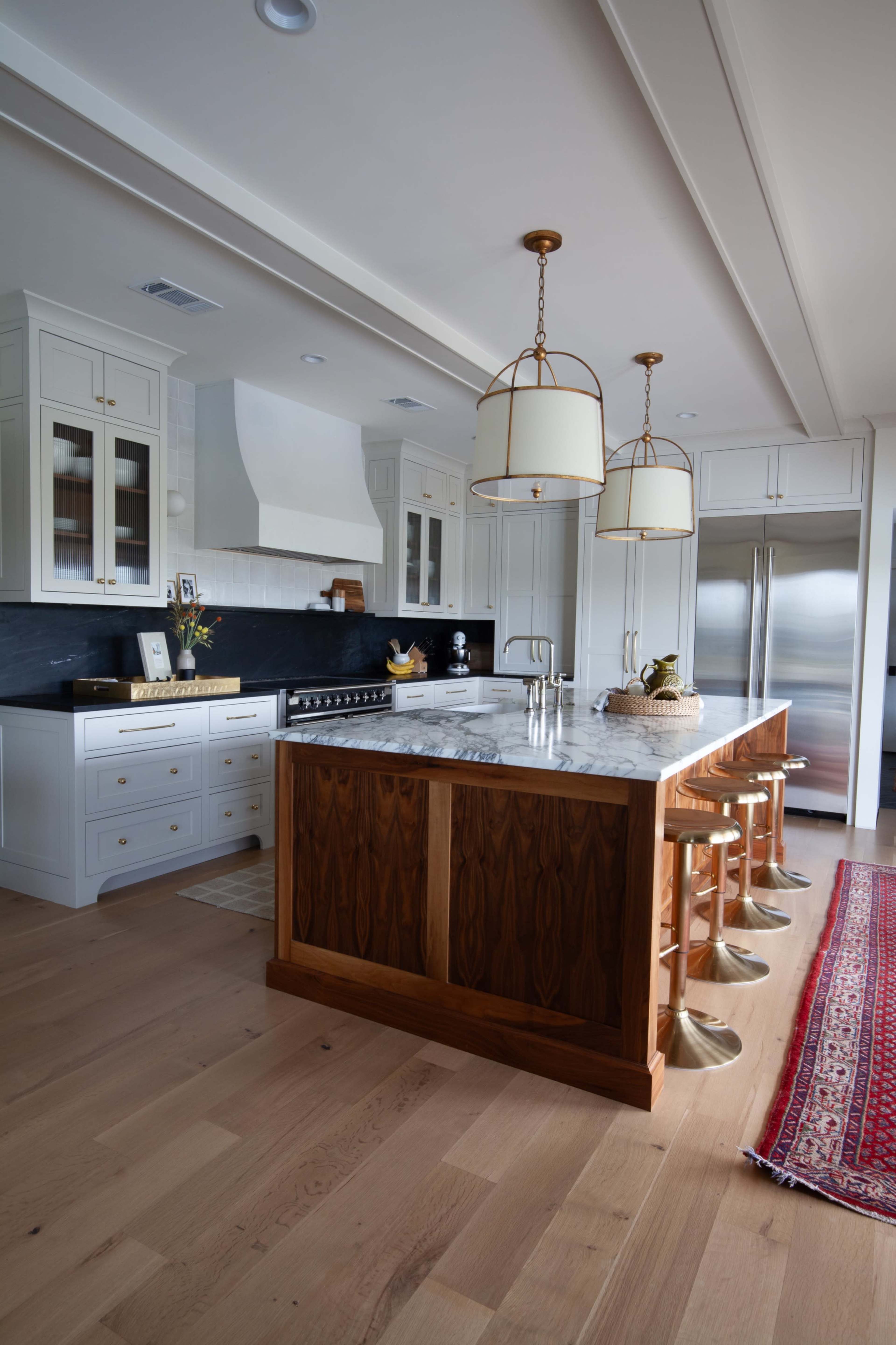A modern kitchen features a central island with a marble countertop, wooden cabinetry, and gold pendant lights, complemented by white cabinets and stainless steel appliances.