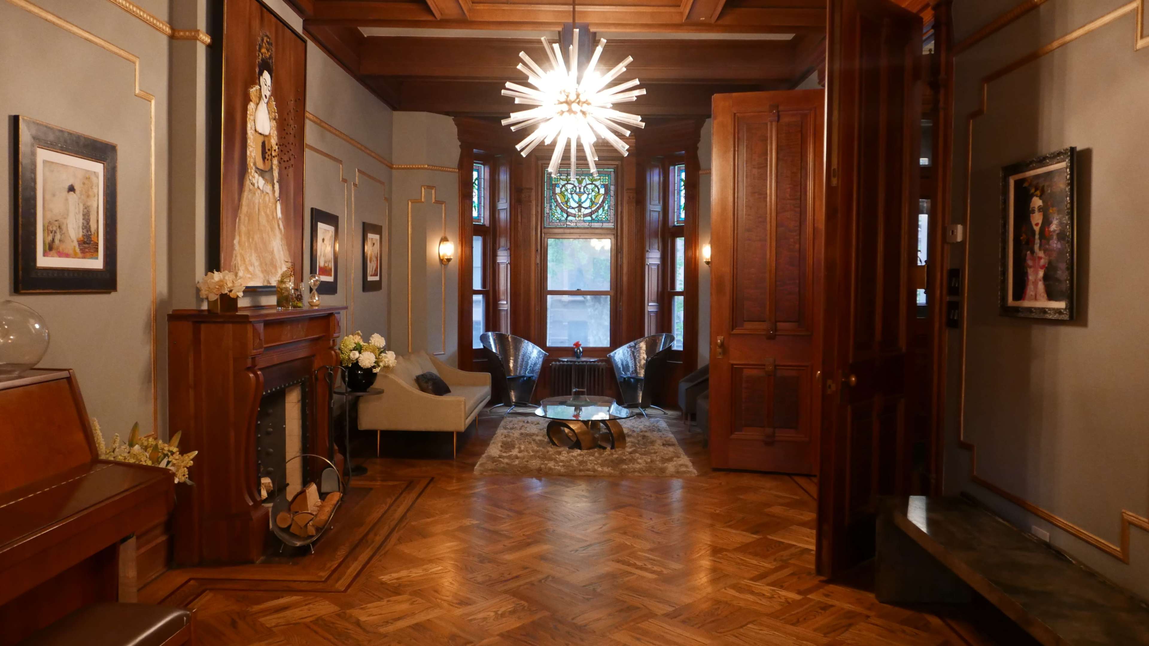 The image shows a stylish interior featuring a wooden floor, a chandelier, a decorative fireplace, and various pieces of furniture arranged in a well-lit room.