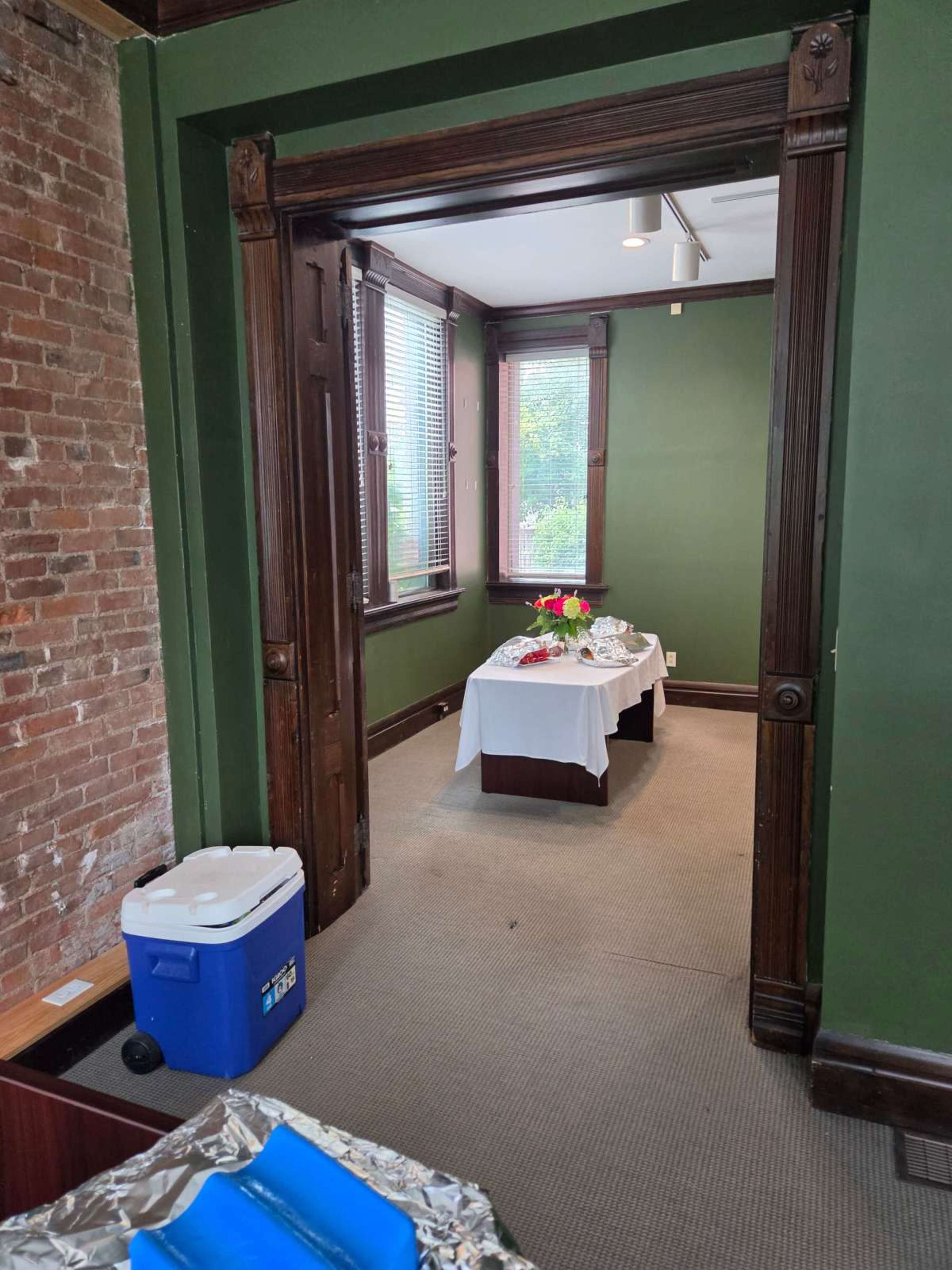 The image shows a doorway leading into a room with a table set up for an event, featuring flowers and a cooler visible in the foreground.