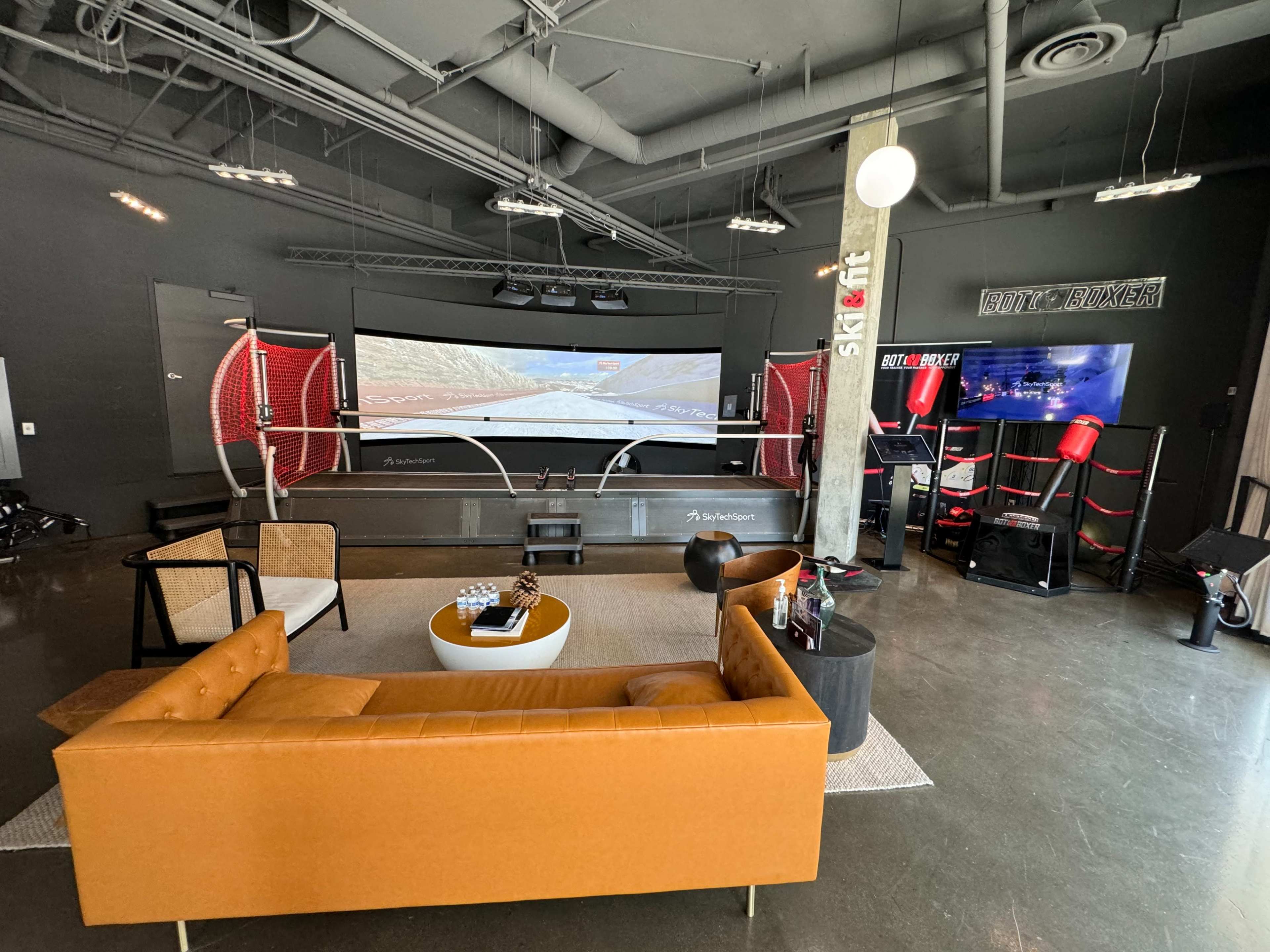 The image shows a modern gaming lounge featuring a large curved screen, racing simulation equipment, and a stylish seating area with a brown sofa and a coffee table.