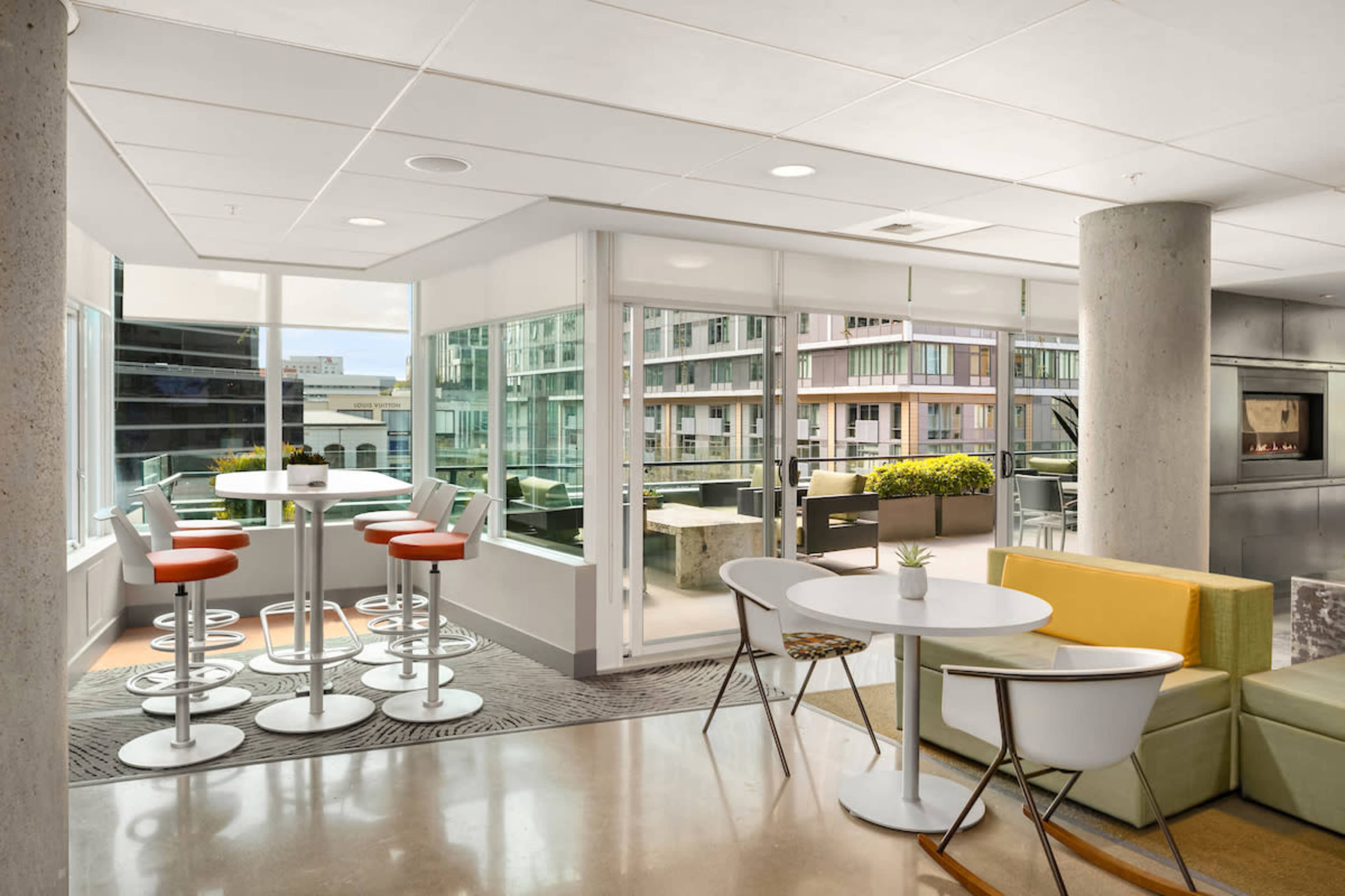 The image shows a modern office lounge area with seating arrangements, large windows providing natural light, and tables surrounded by stools.