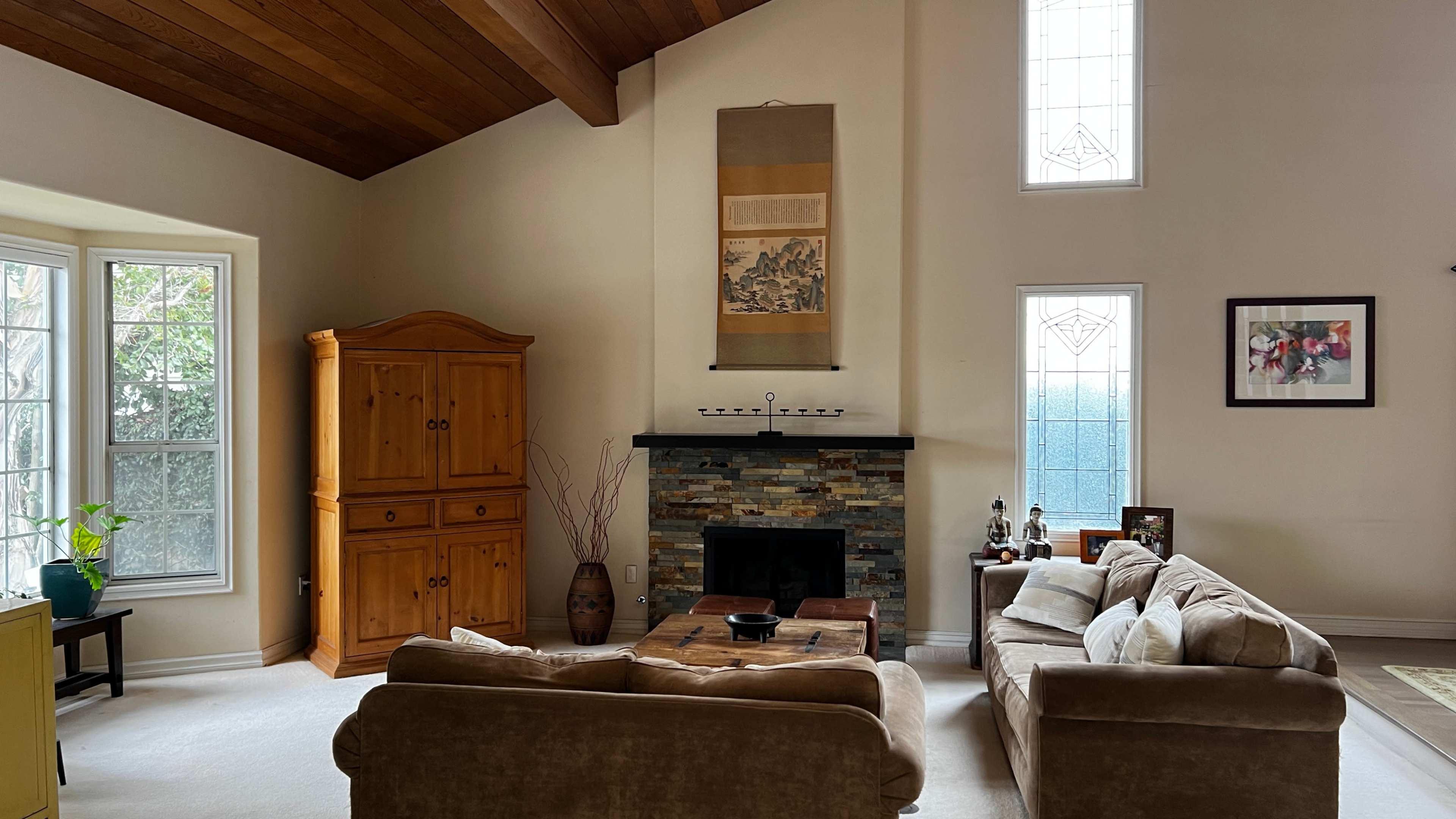 A living room features a stone fireplace, large windows, a wooden cabinet, and comfortable seating arranged around a coffee table.