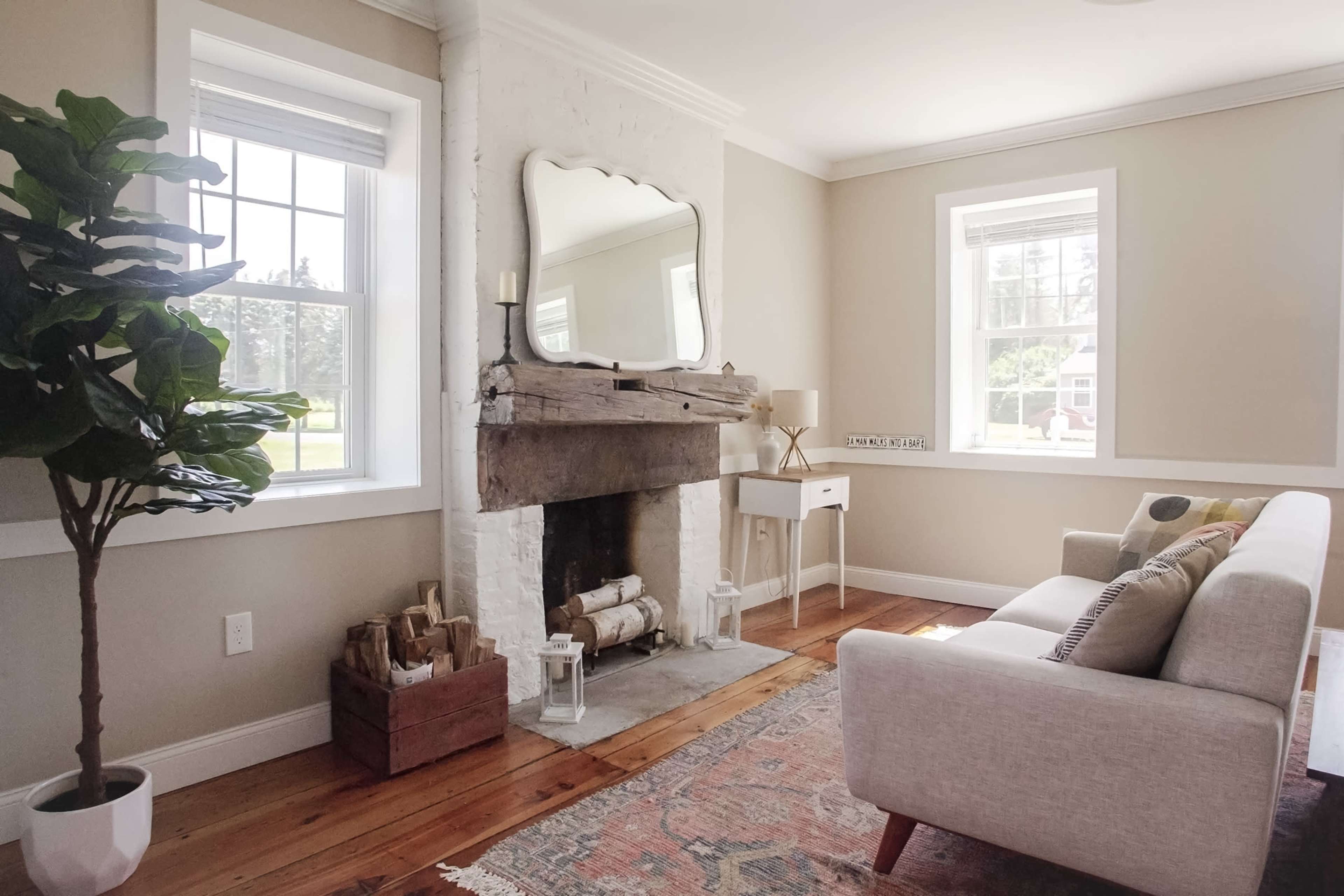 A cozy living room features a white brick fireplace with a wooden mantel, a large mirror above it, and a sofa facing two windows.