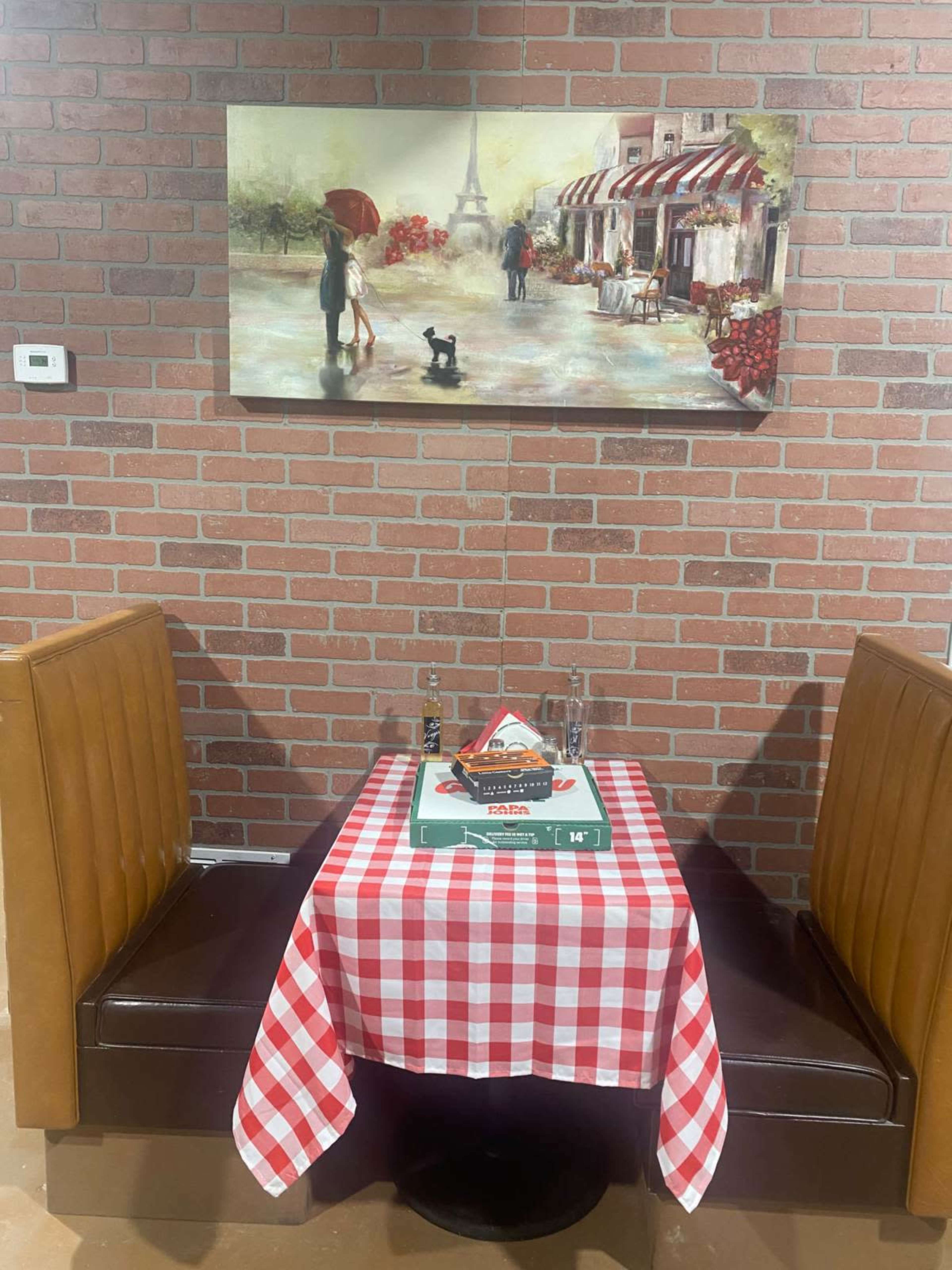 A small dining area features a checkered tablecloth, a pizza box, and a painting of a Parisian street scene on the brick wall.